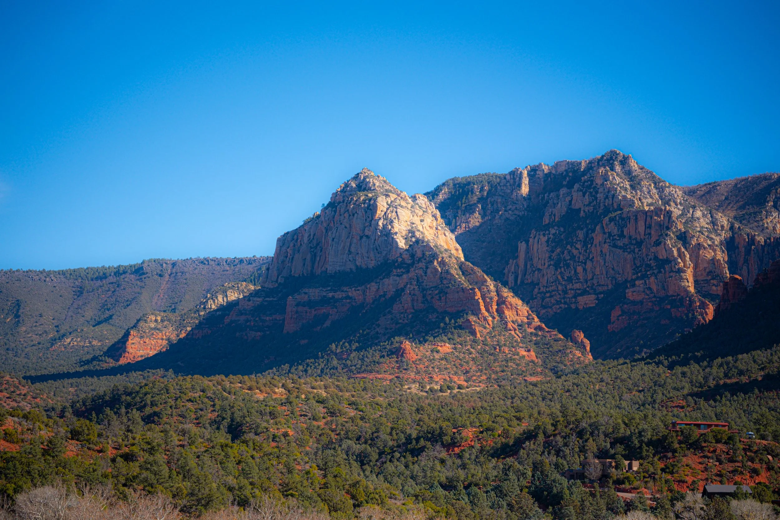 A landscape of mountain peaks with reddish and beige rock formations, green forested slopes, clear blue sky, and a few houses near the bottom right.