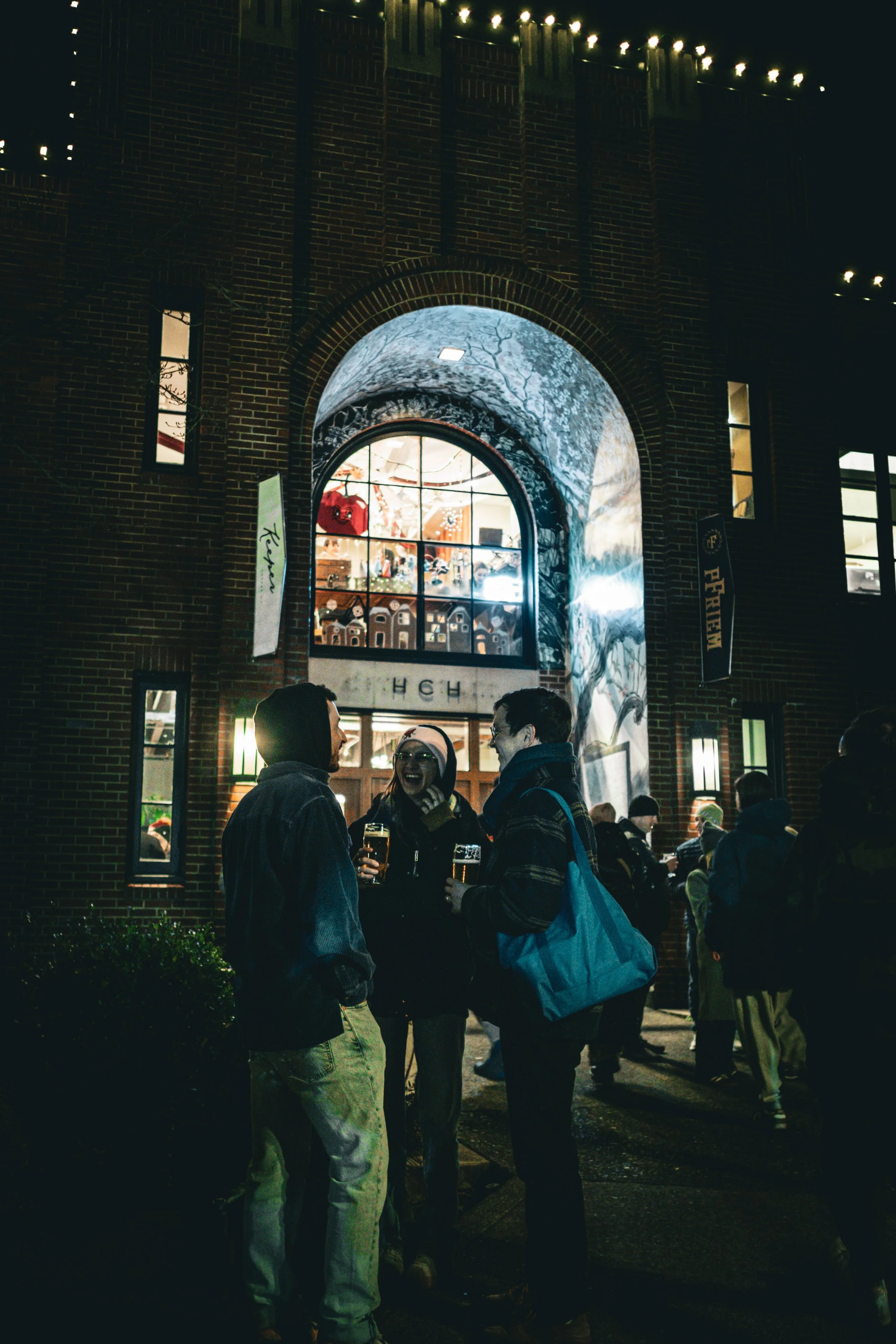 People gathered outside a brick building at night, socializing and holding drinks, with illuminated windows and string lights overhead.