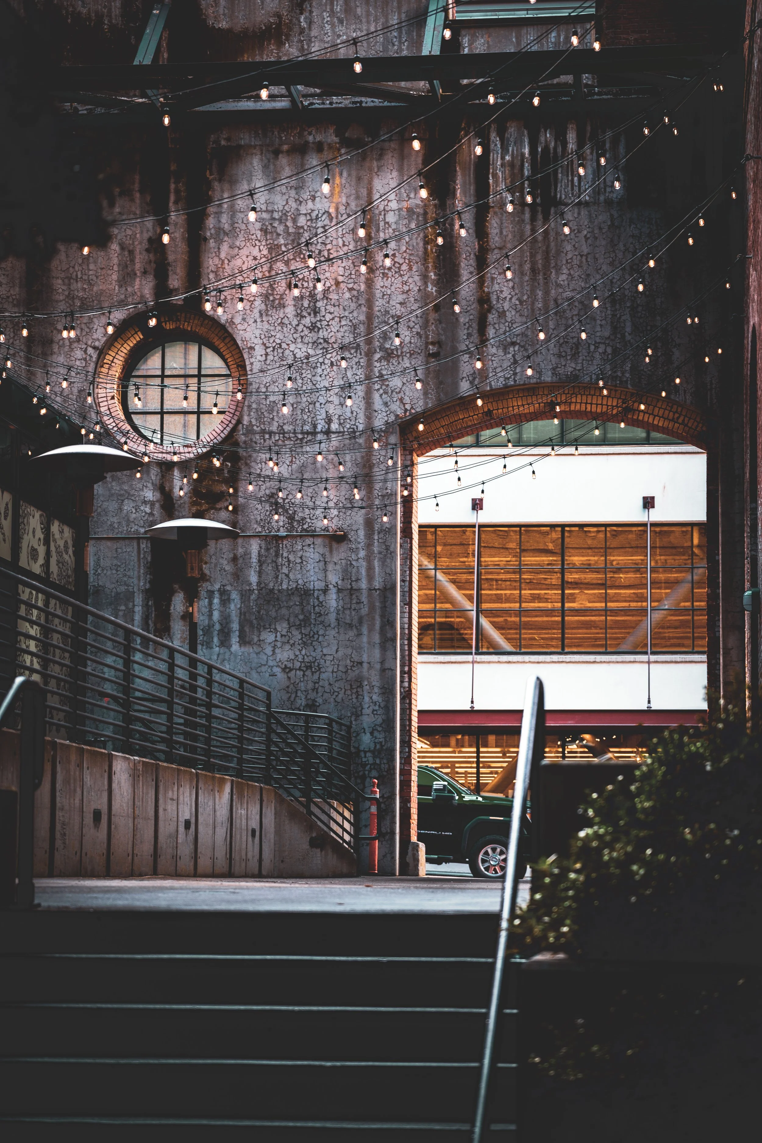 An urban outdoor patio area with string lights hanging above, a textured weathered wall with a round window, and a parking lot visible through an archway with a black truck parked nearby.