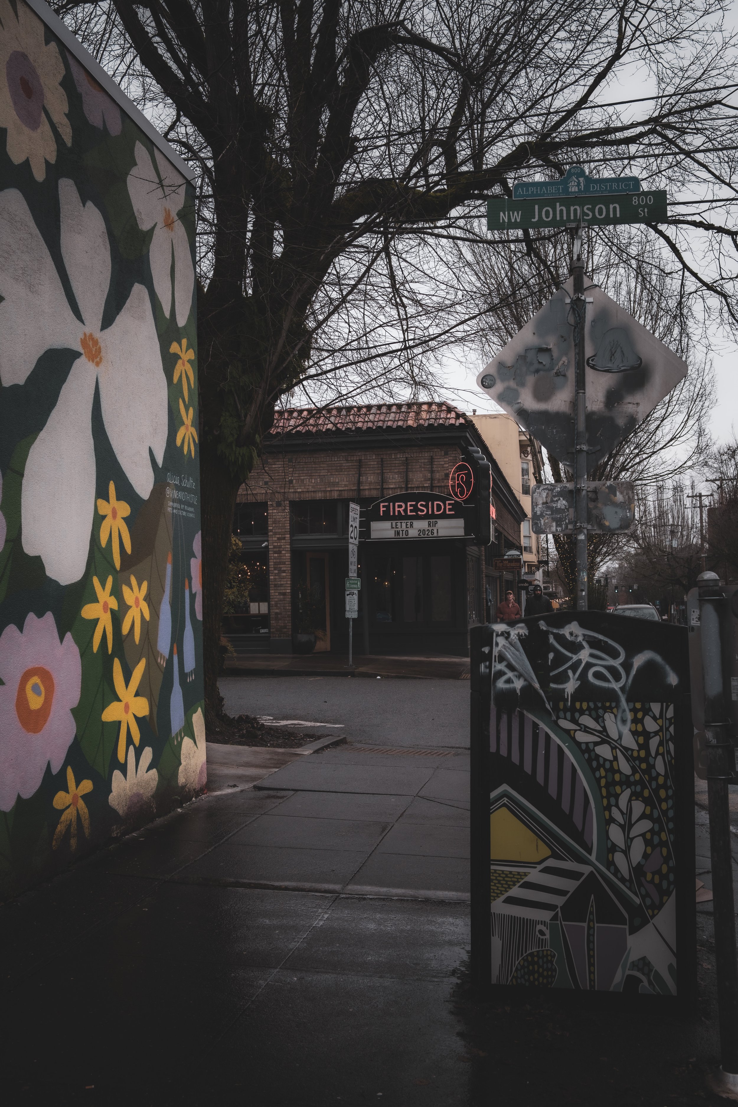 A street scene showing a mural on the left, street signs above, a brick building with a neon sign that reads 'FIRESIDE,' and some trees in the background. The sidewalk appears wet.
