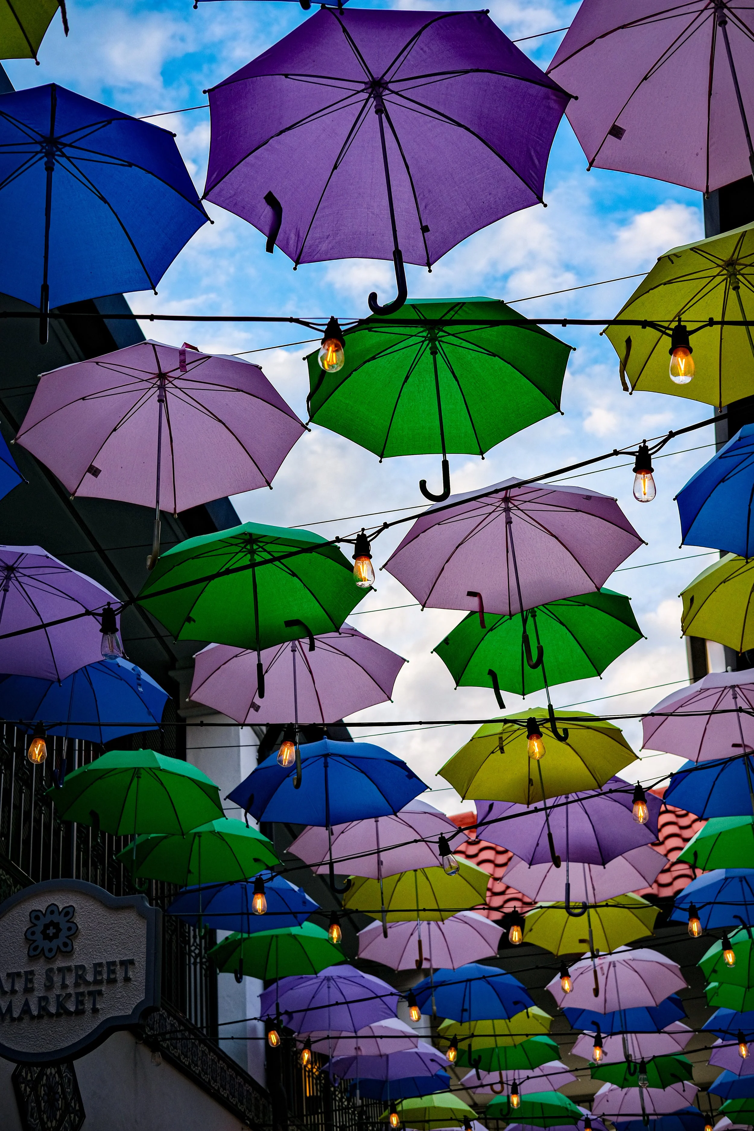Colorful umbrellas hung overhead at an outdoor market with string lights and a blue sky.