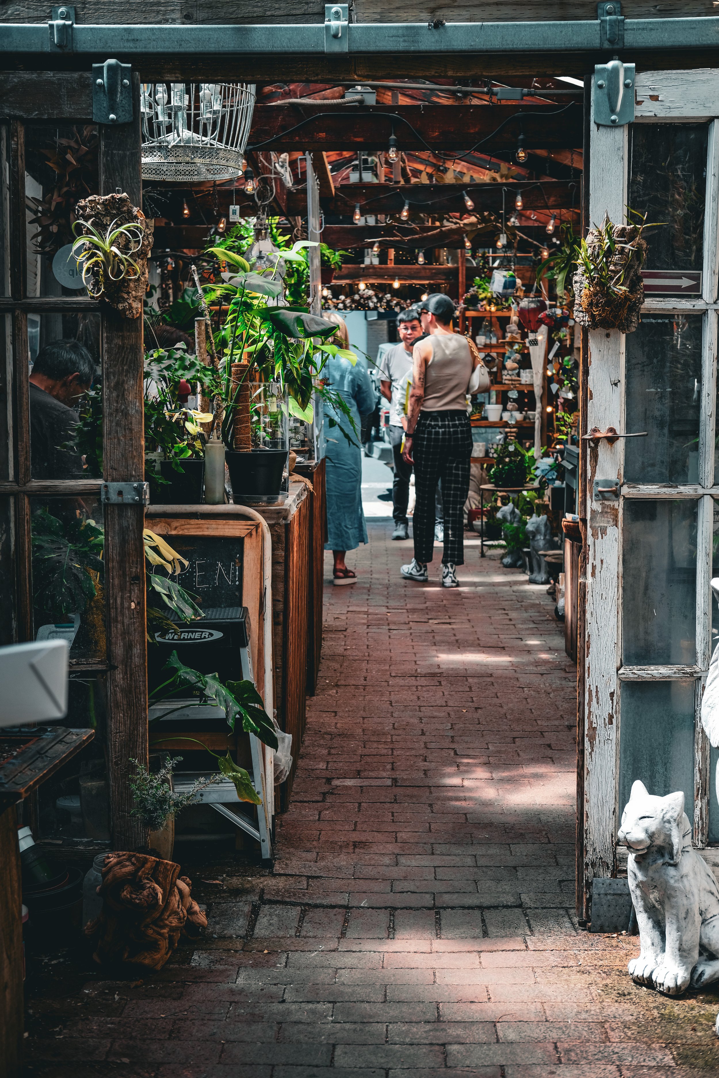 People browsing a rustic outdoor marketplace or garden shop, with plants, decorative items, and a white cat statue near the entrance.