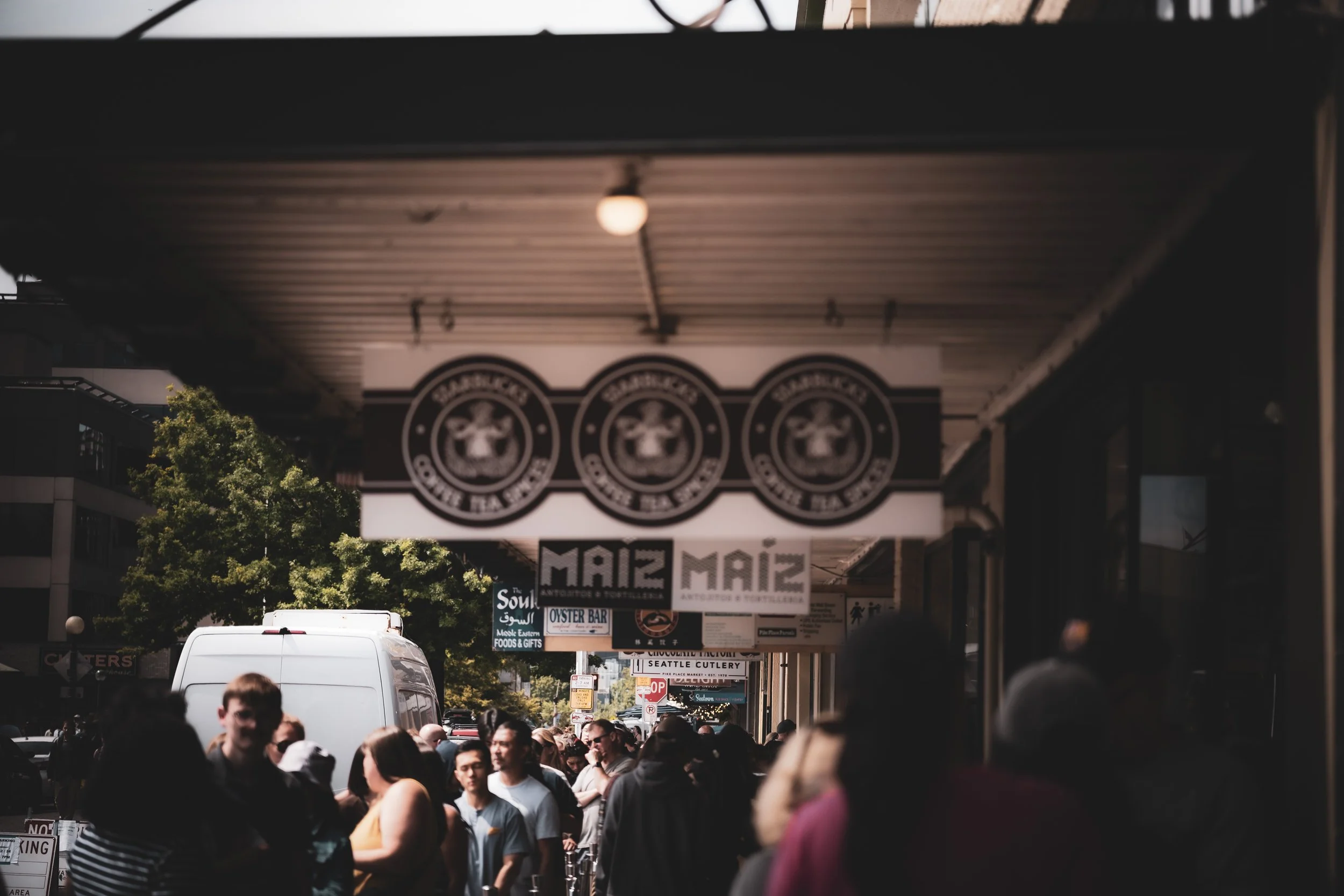 Crowd of people walking along city sidewalk shaded by storefront awning with hanging signs, and a white van parked on the street in the background.