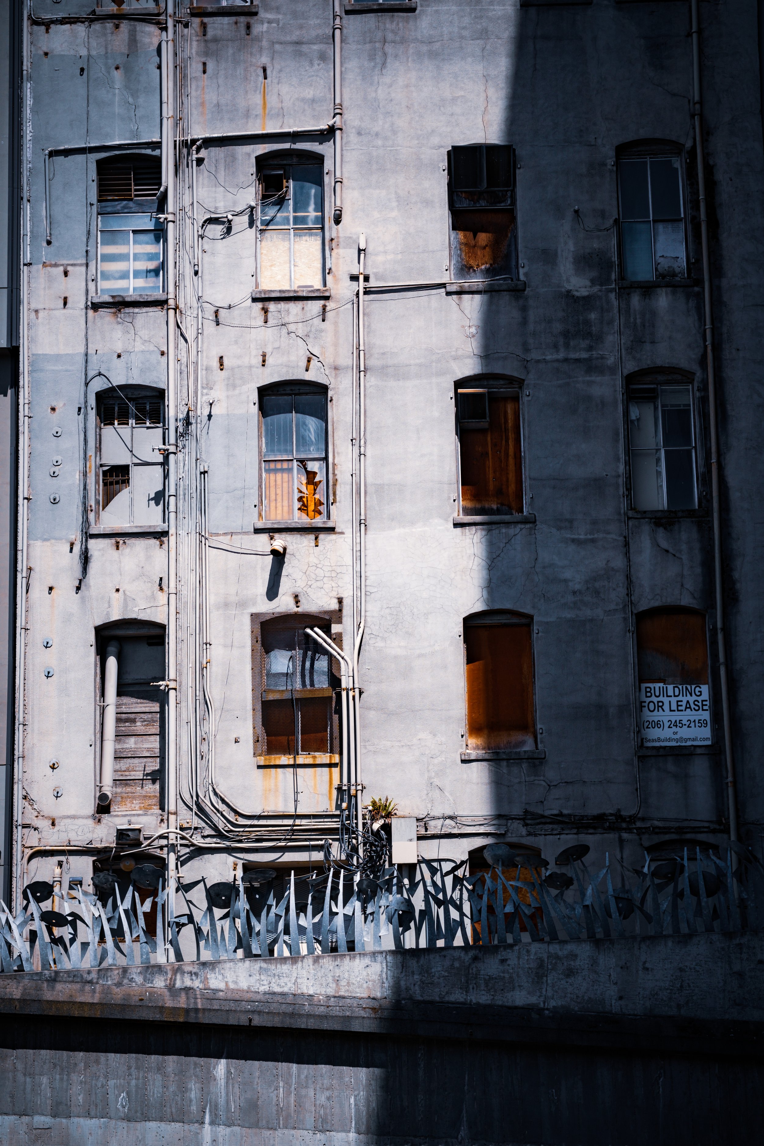 A weathered, gray building with cracked walls and multiple broken or boarded-up windows, some with curtains visible inside. There are numerous pipes and cables running along the exterior. A sign on one window indicates the building is available for l