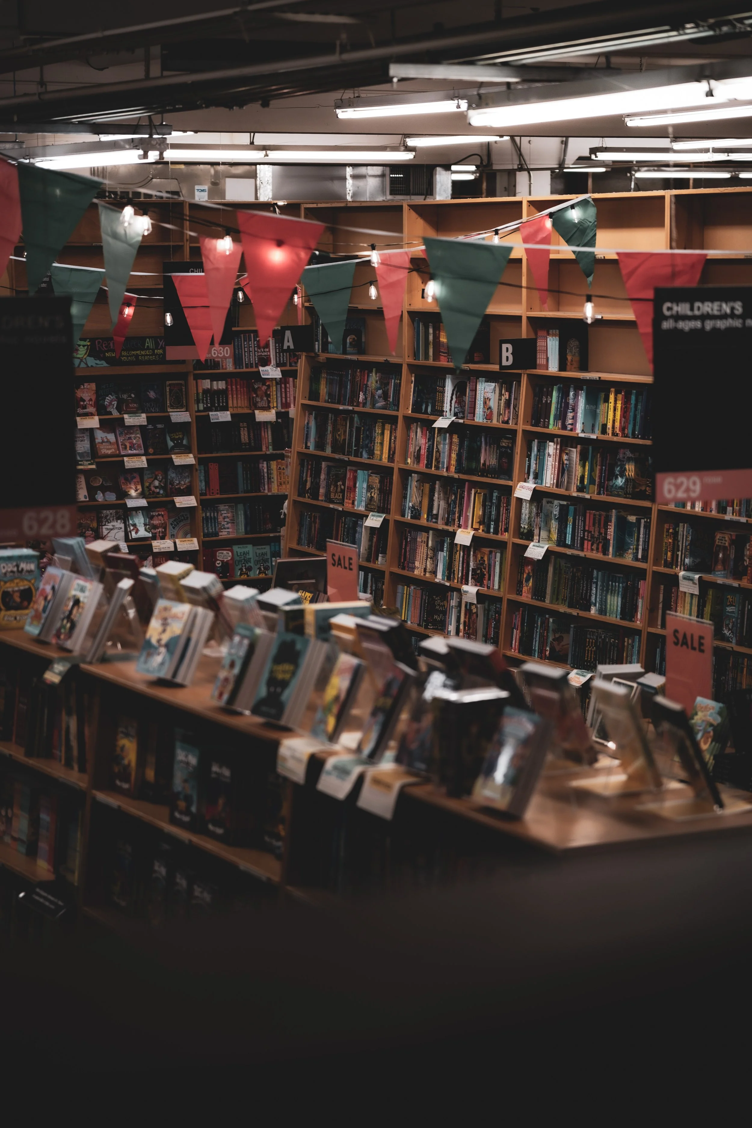 Bookshelves filled with books in a bookstore, decorated with triangular red and green pennant banners and string lights.