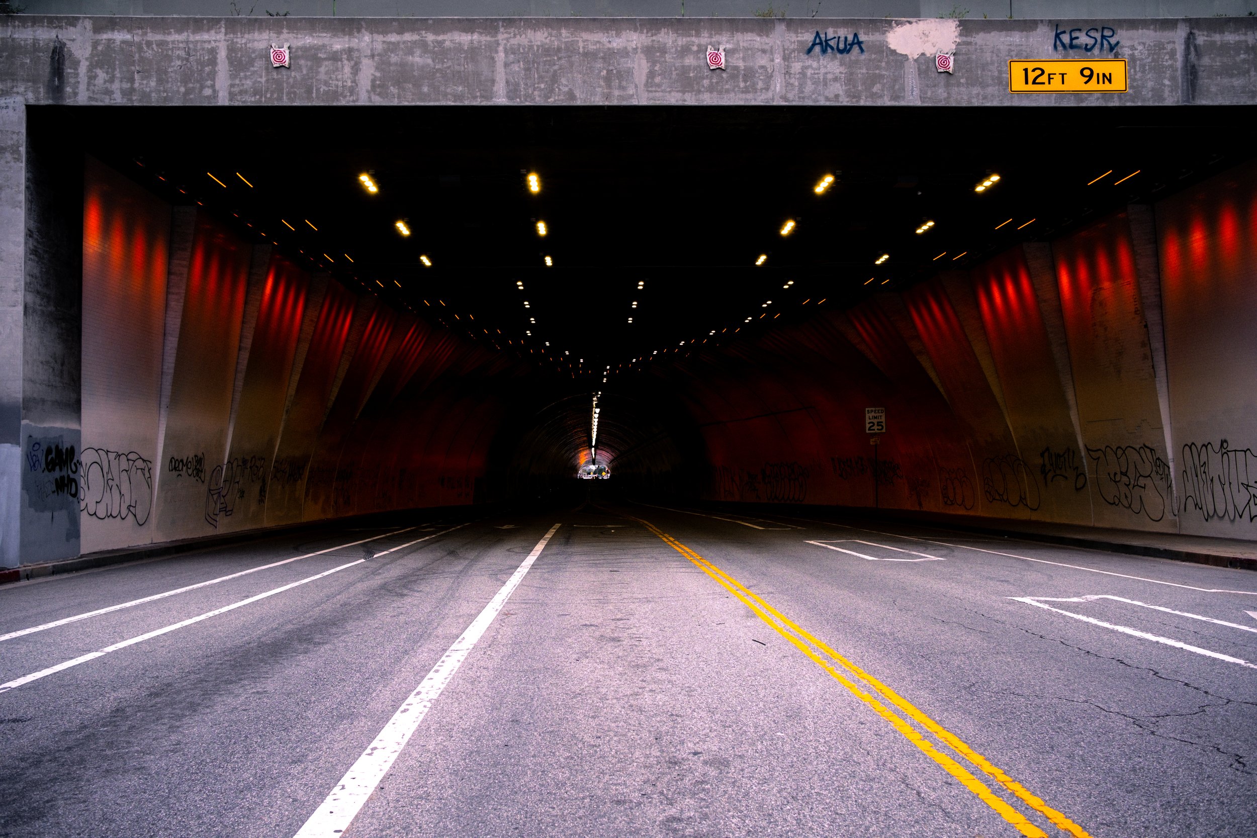 View of a tunnel on a city street with graffiti on the walls, illuminated by ceiling lights, with a sign indicating a height clearance of 12 feet 9 inches.