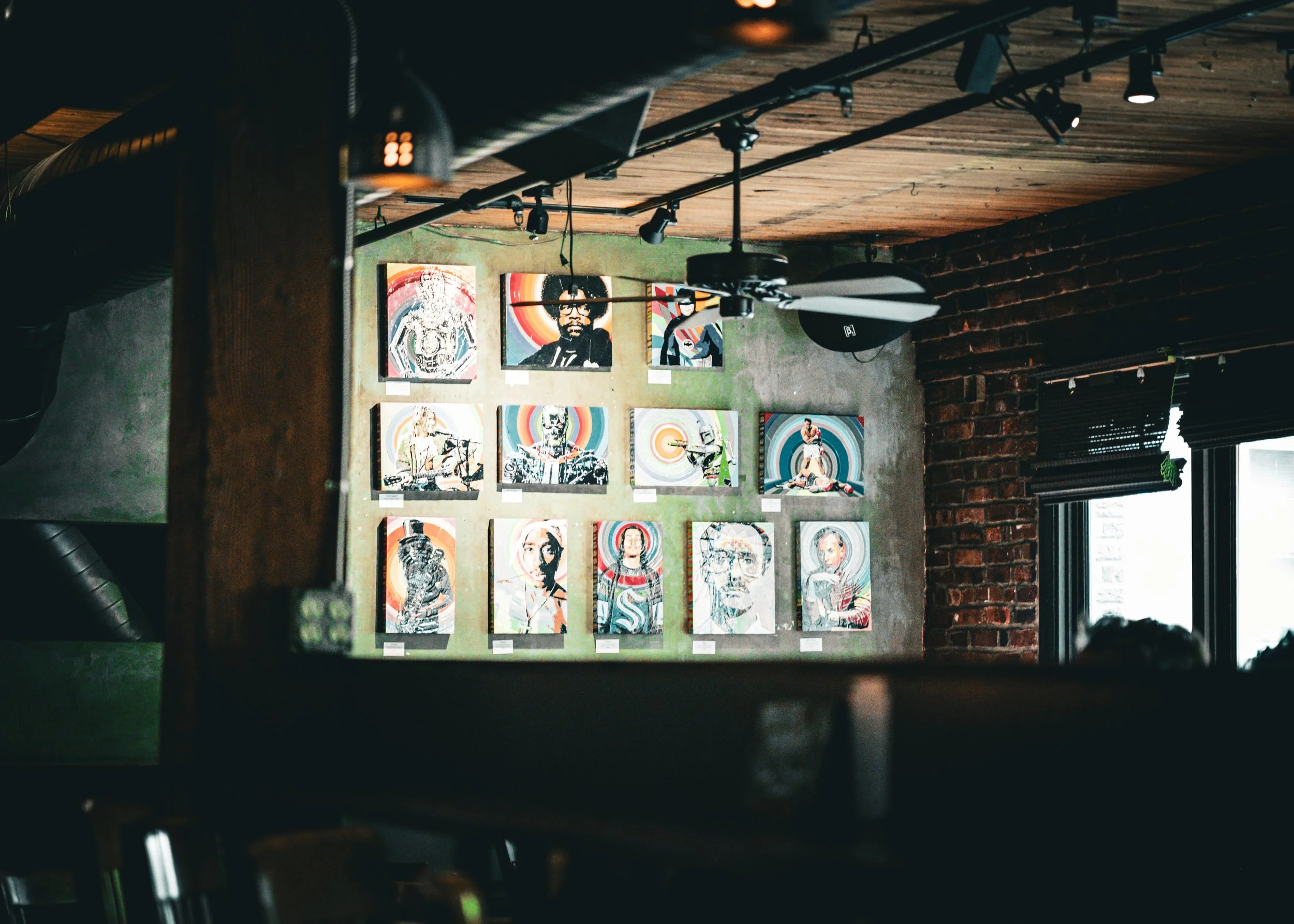 Art gallery with colorful portraits of people on the wall, ceiling fan, large window with blinds, exposed brick wall, and track lighting.