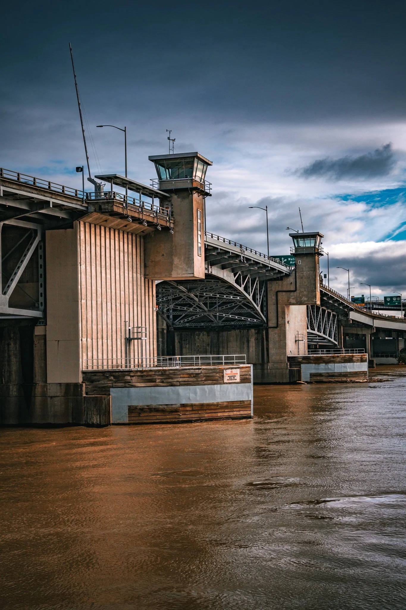 A bridge with two control towers over a brown river, with cloudy sky in the background. Portland Bridge. 
