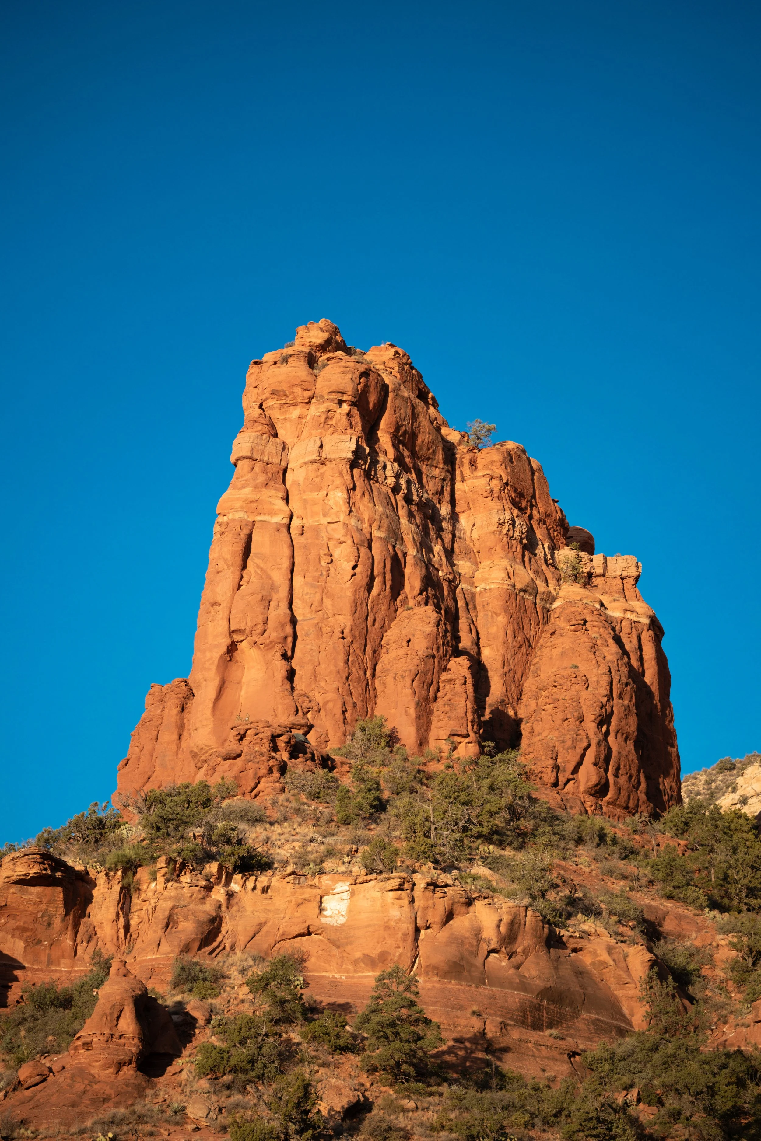 A tall red sandstone formation against a blue sky with sparse green vegetation at its base.