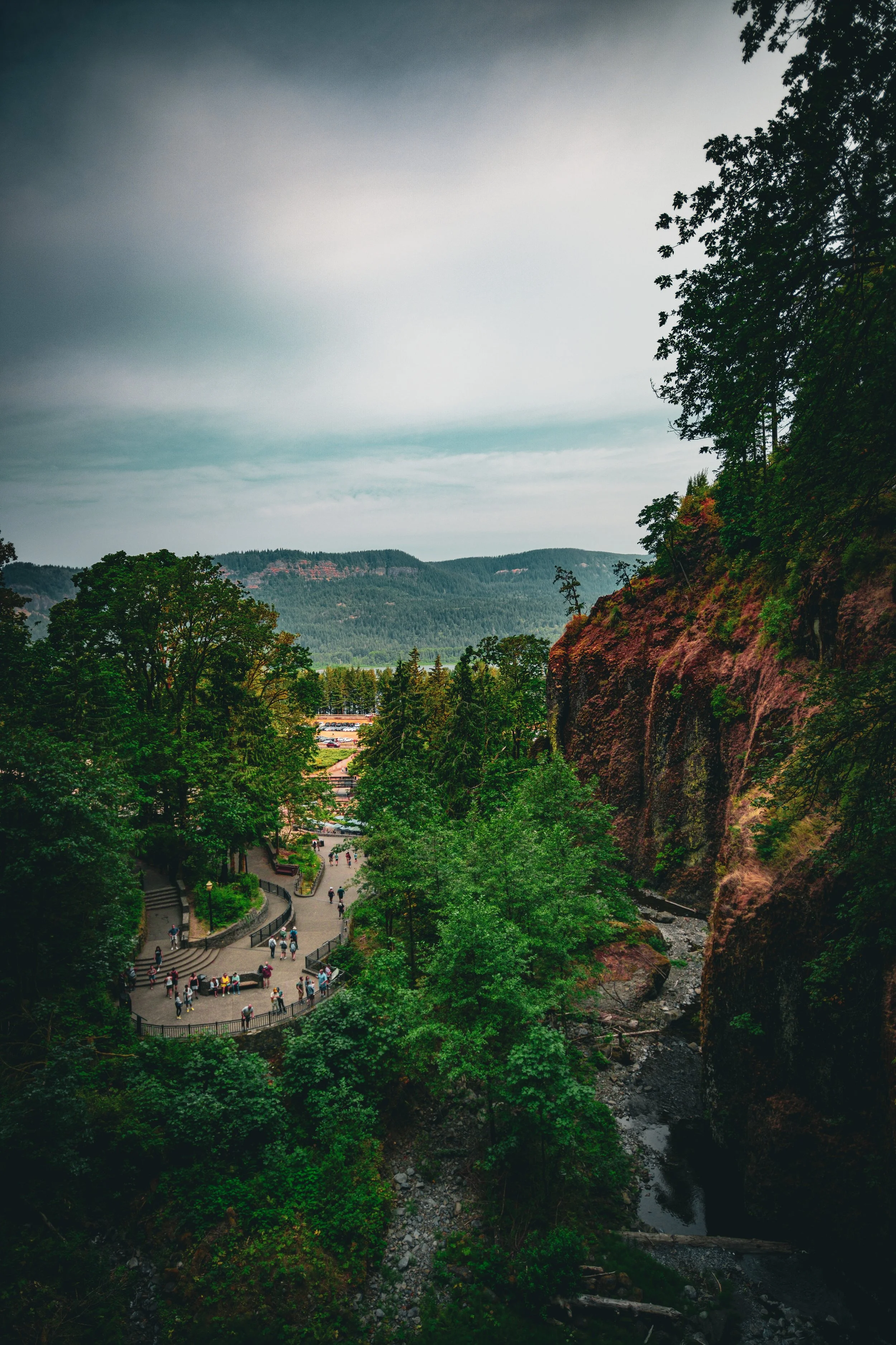 A scenic view of a park with walking paths filled with people, surrounded by green trees and red rocky cliffs, with forested hills and mountains in the background under a cloudy sky.