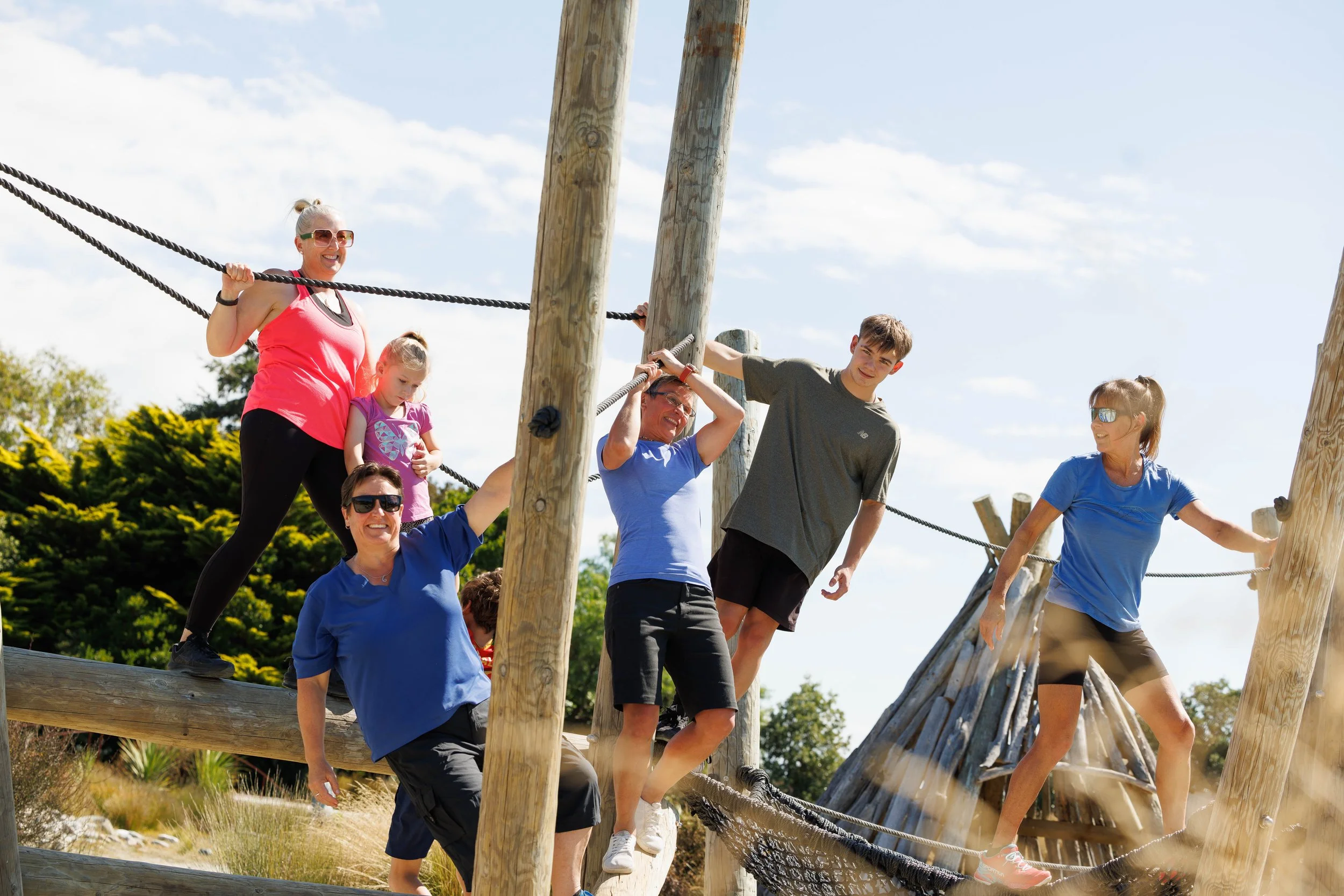 People making their way across a wooden balance beam in an outdoor ropes course on a sunny day.