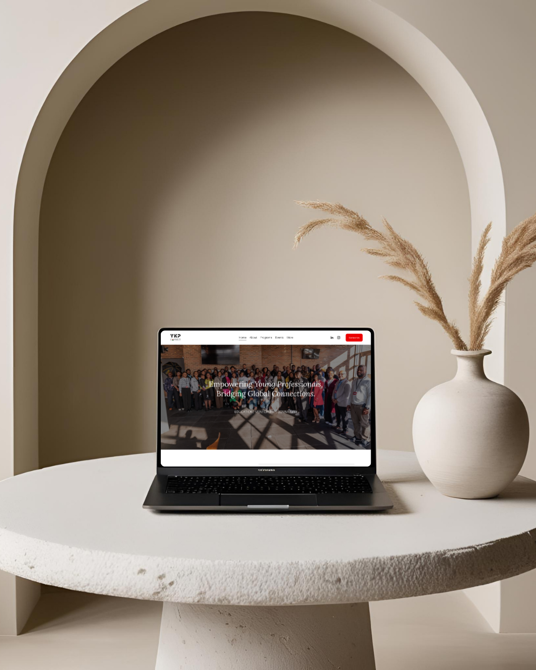 A laptop on a white round table displaying a website titled 'Empowering Young Professionals, Bridging Global Connections.' Next to the laptop is a beige vase with dried pampas grass, set inside an arched architectural opening with beige walls.