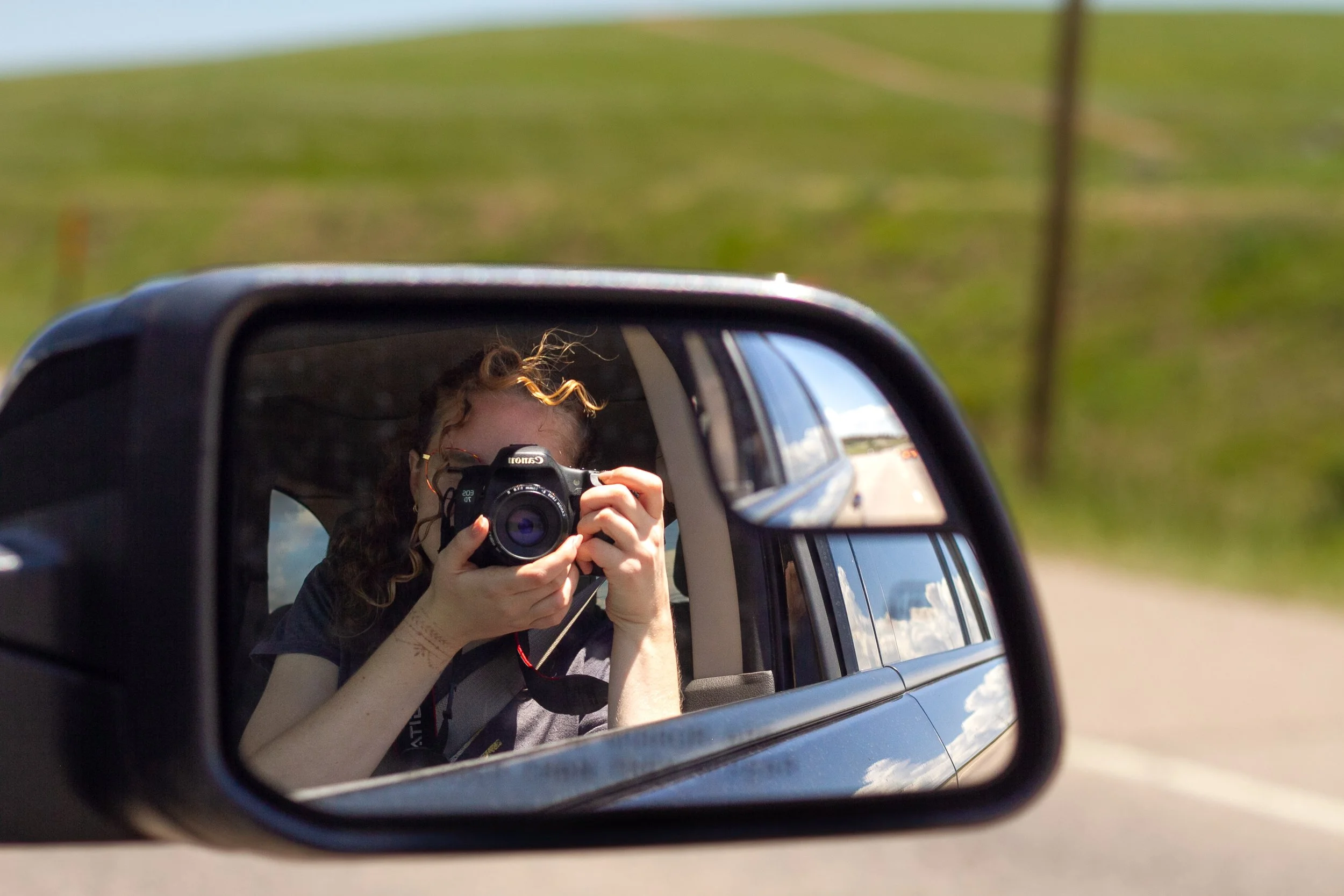 Person taking a photo with a Canon camera in a car's side mirror, with a grassy landscape and blue sky reflected in the mirror.