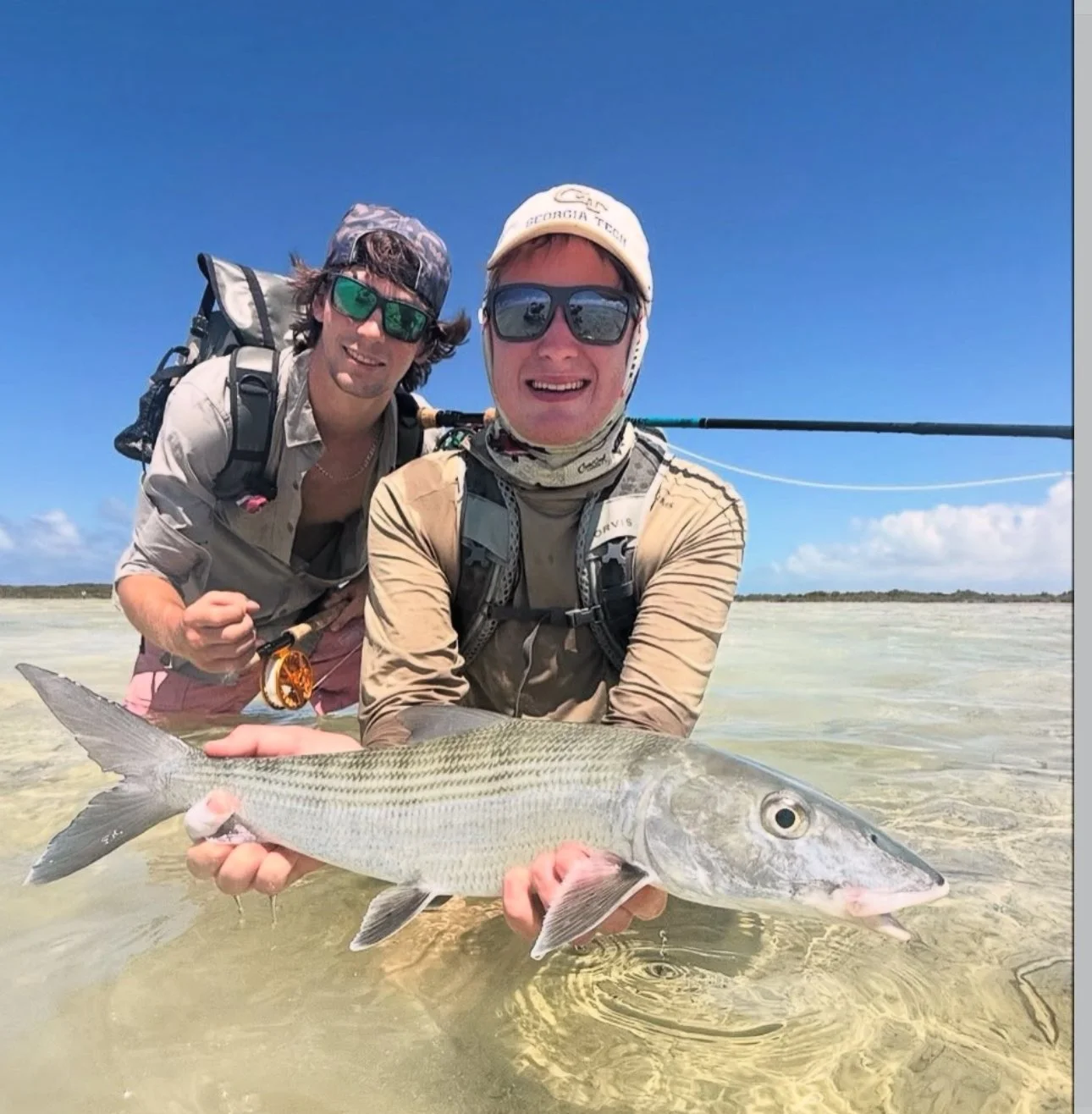 Two men in fishing gear standing in shallow water at the beach, holding a large bonefish with sandy shore and blue sky in the background.