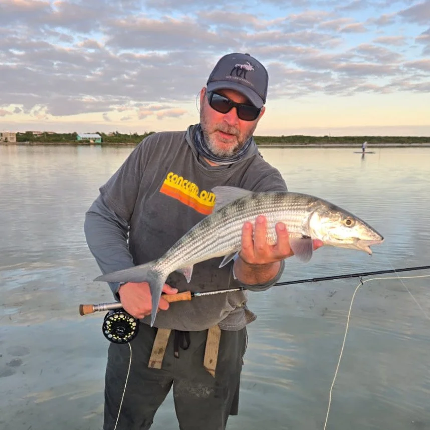 Man in dark sunglasses and a baseball cap holding a large bonefish with a fishing rod, standing in shallow water during sunset.