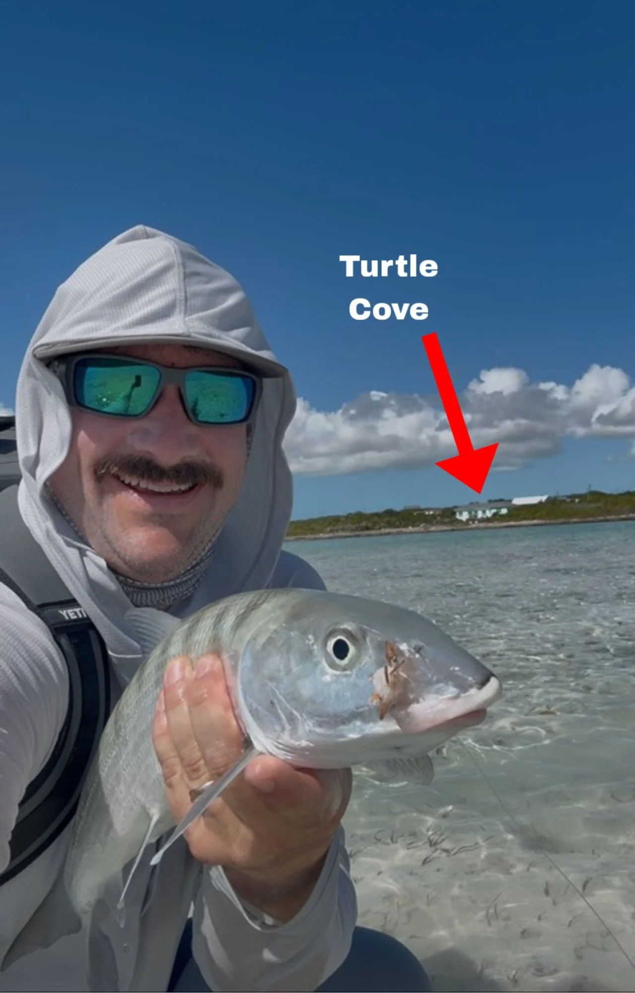 A man wearing sunglasses and a sun protection hood takes a selfie while holding a fish he caught. In the background, a body of water and a shoreline is visible, with a small building labeled 'Turtle Cove' on it.