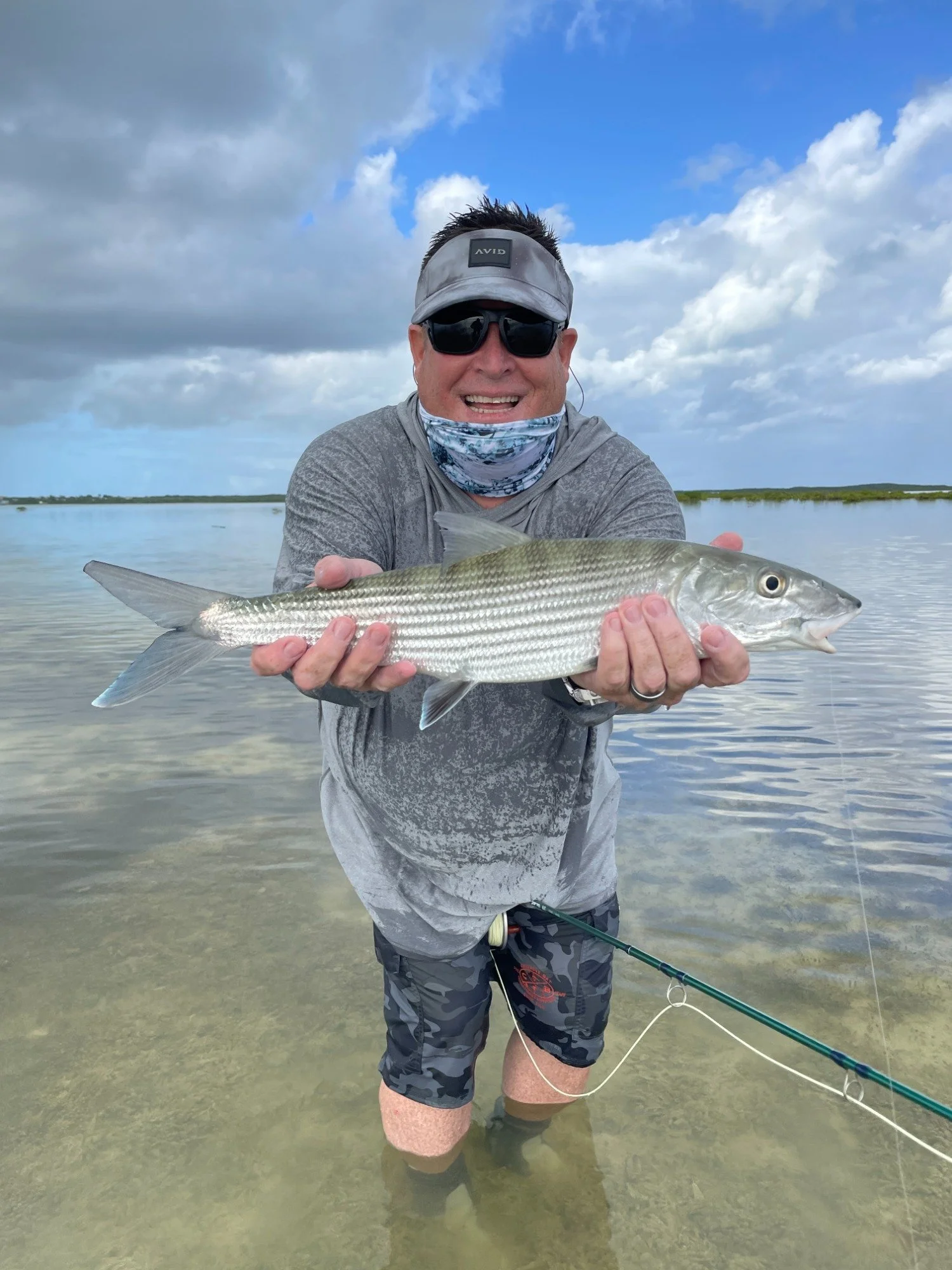 Man wearing sunglasses, hat, and face mask holding a large bonefish in shallow water with cloudy sky in background.