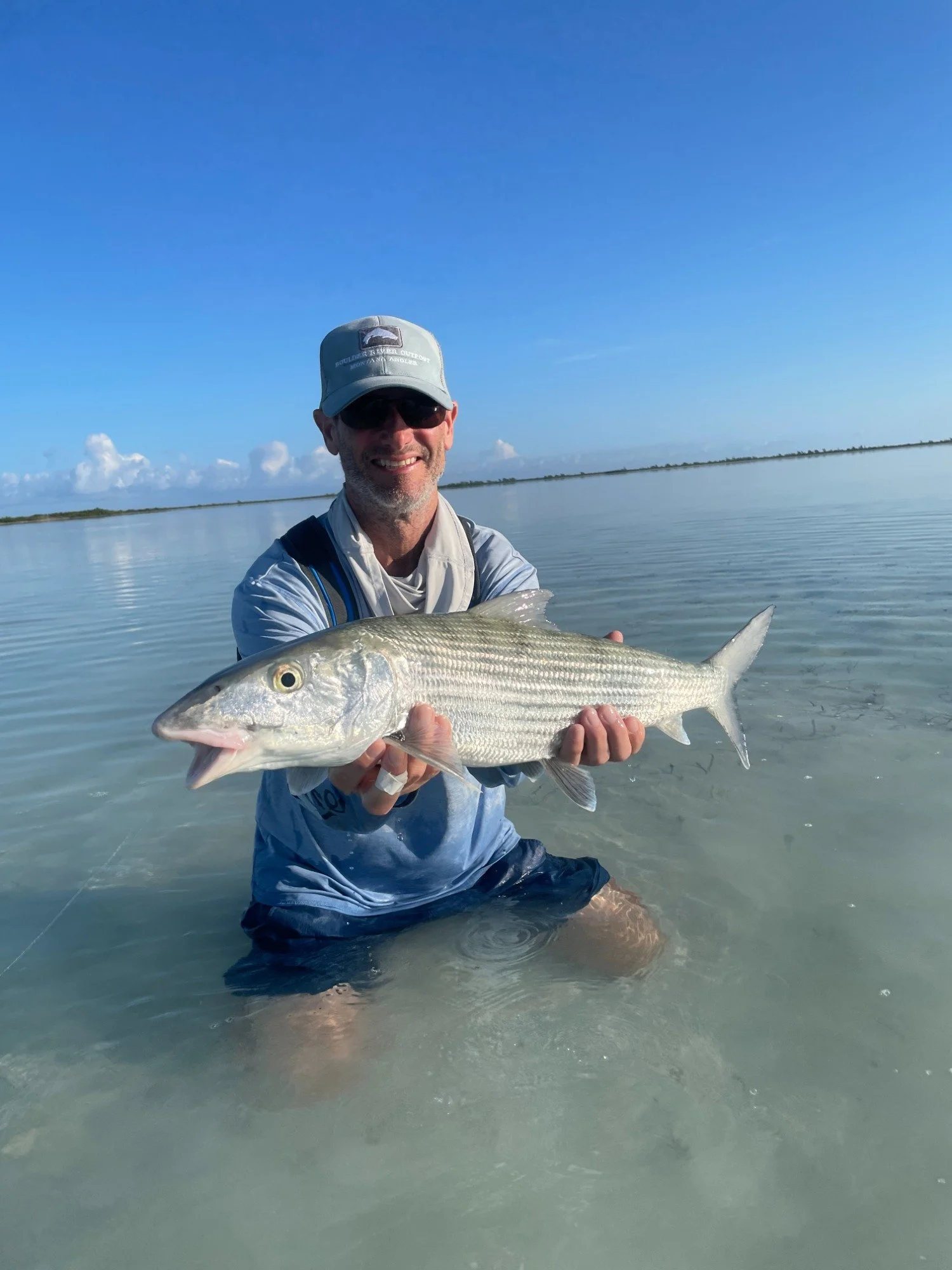 Man standing waist-deep in water, wearing sunglasses and a hat, holding a large bonefish, with a calm body of water and blue sky in the background.
