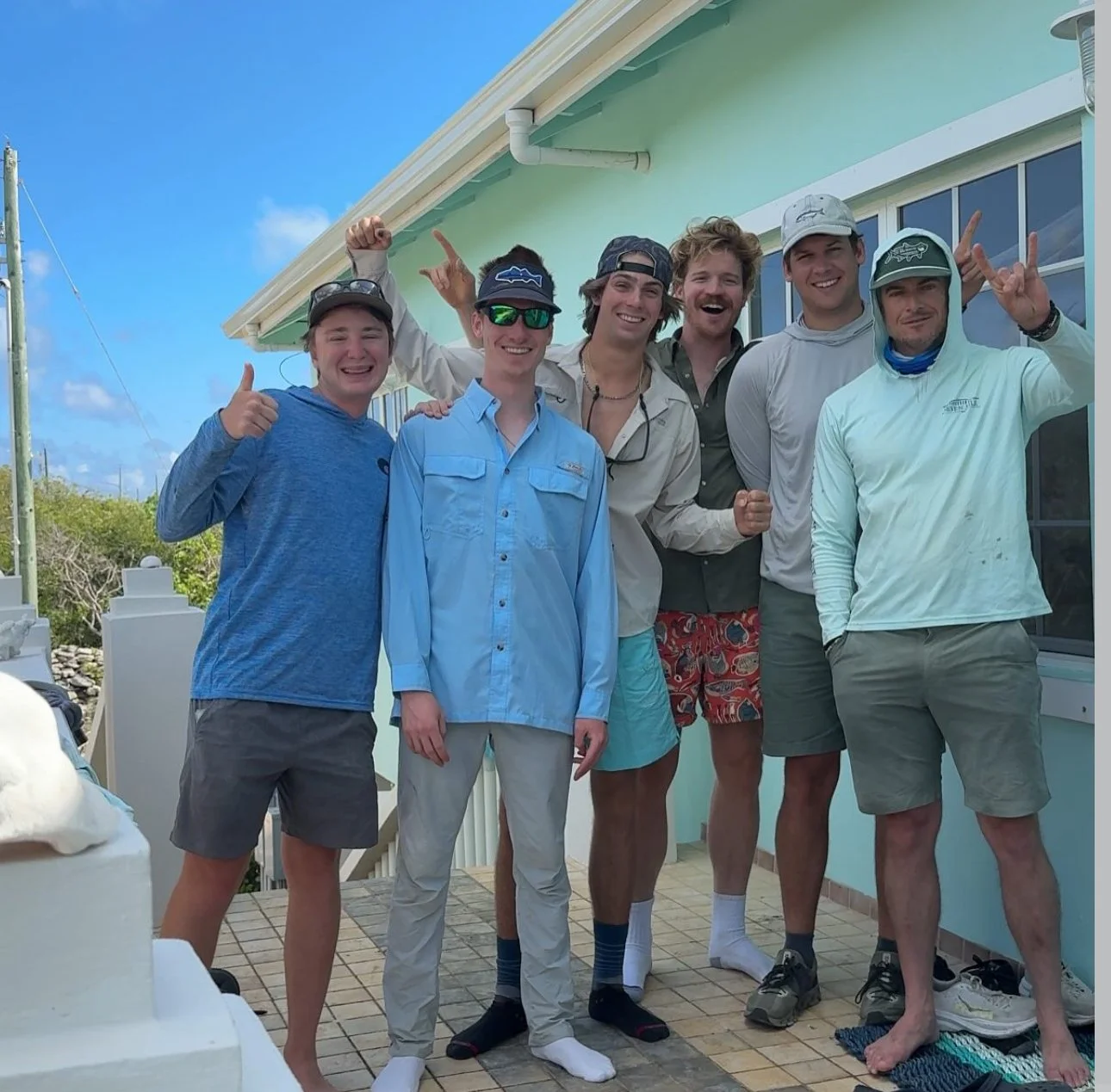 A group of six young men standing on a balcony outside a house, posing and smiling for a photo. They are dressed casually, some wearing hats and sunglasses, with one giving a thumbs-up and another making a shaka sign. The background shows a clear sky