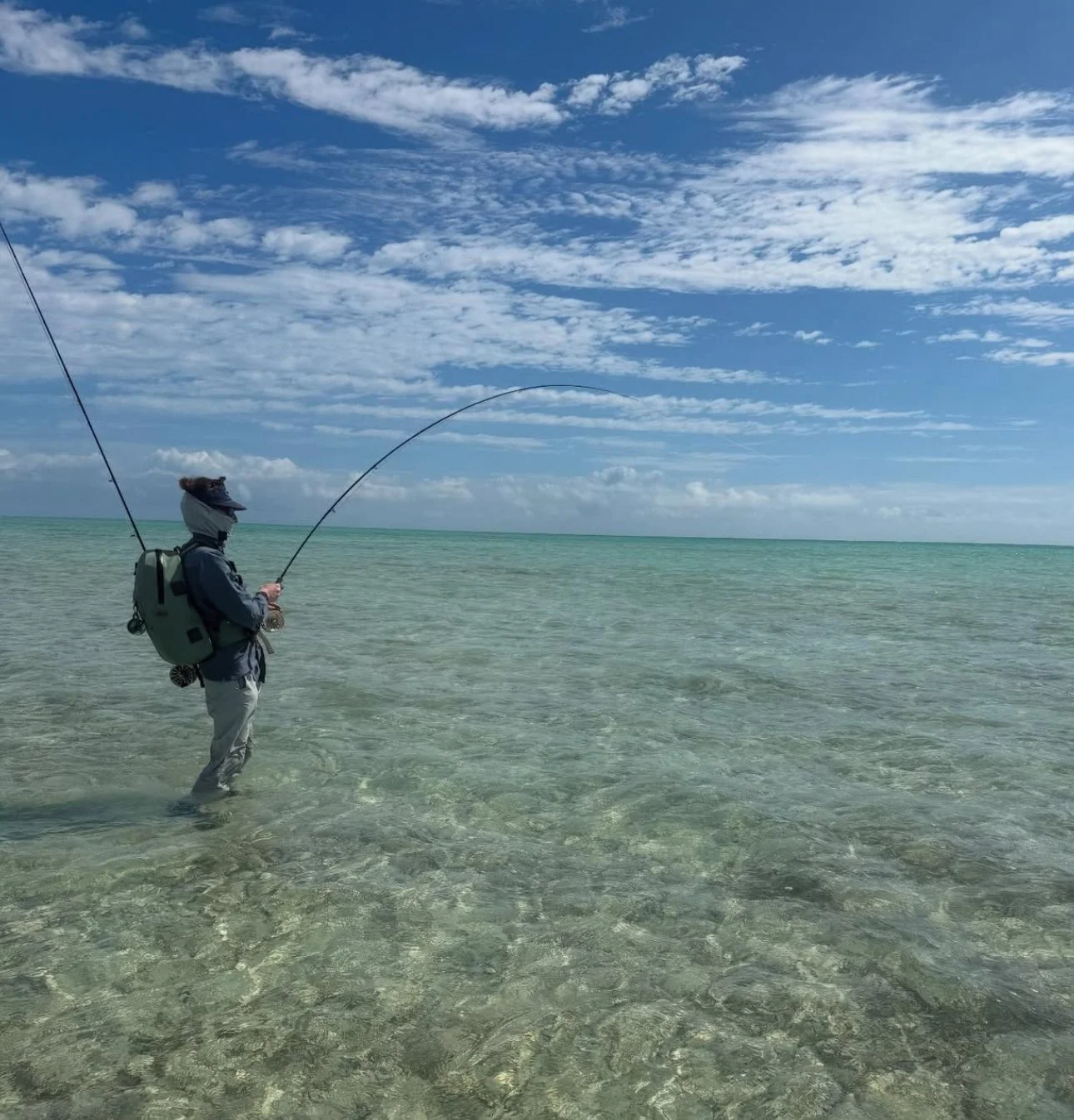 A person bonefishing in shallow, clear water near the beach under a partly cloudy sky.