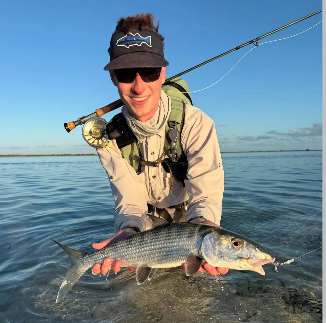 A smiling man wearing sunglasses, a hat with a fish logo, and outdoor fishing gear, holding a large bonefish near the water during daytime.