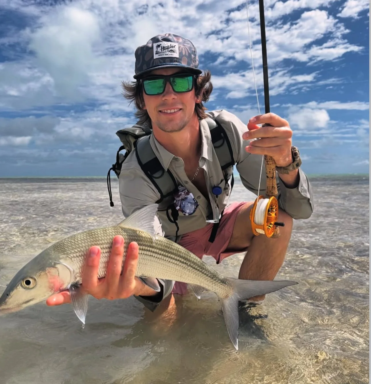 A man kneeling on the beach holding a large bonefish he caught while fishing. He is wearing sunglasses, a hat, a light-colored shirt, and pink shorts, and is smiling at the camera. The background shows a partly cloudy sky and shallow water.