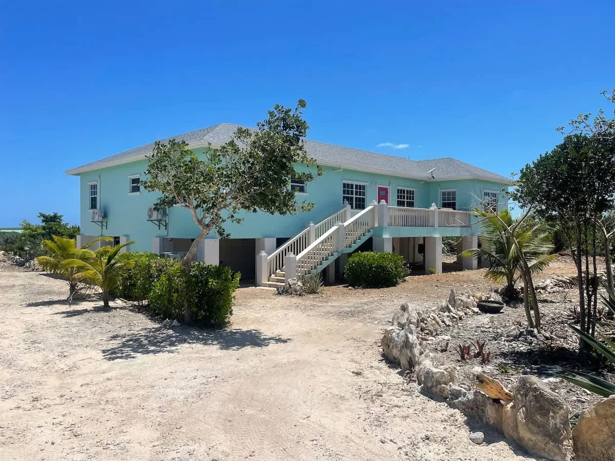 A raised light blue house with a staircase leading to a front porch, surrounded by sparse desert landscaping, trees, and small plants under a clear blue sky.