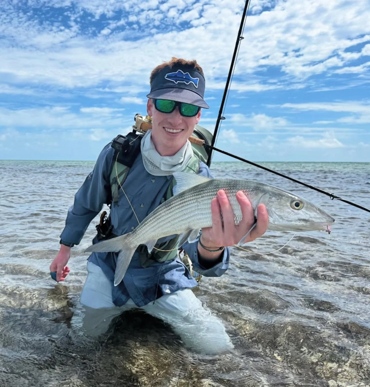 A man in fishing gear holding a large bonefish at the beach with water and sky in the background.