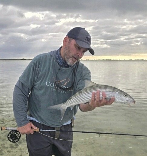 Man holding a bonefish he caught while fishing on a calm body of water, with an overcast sky in the background.