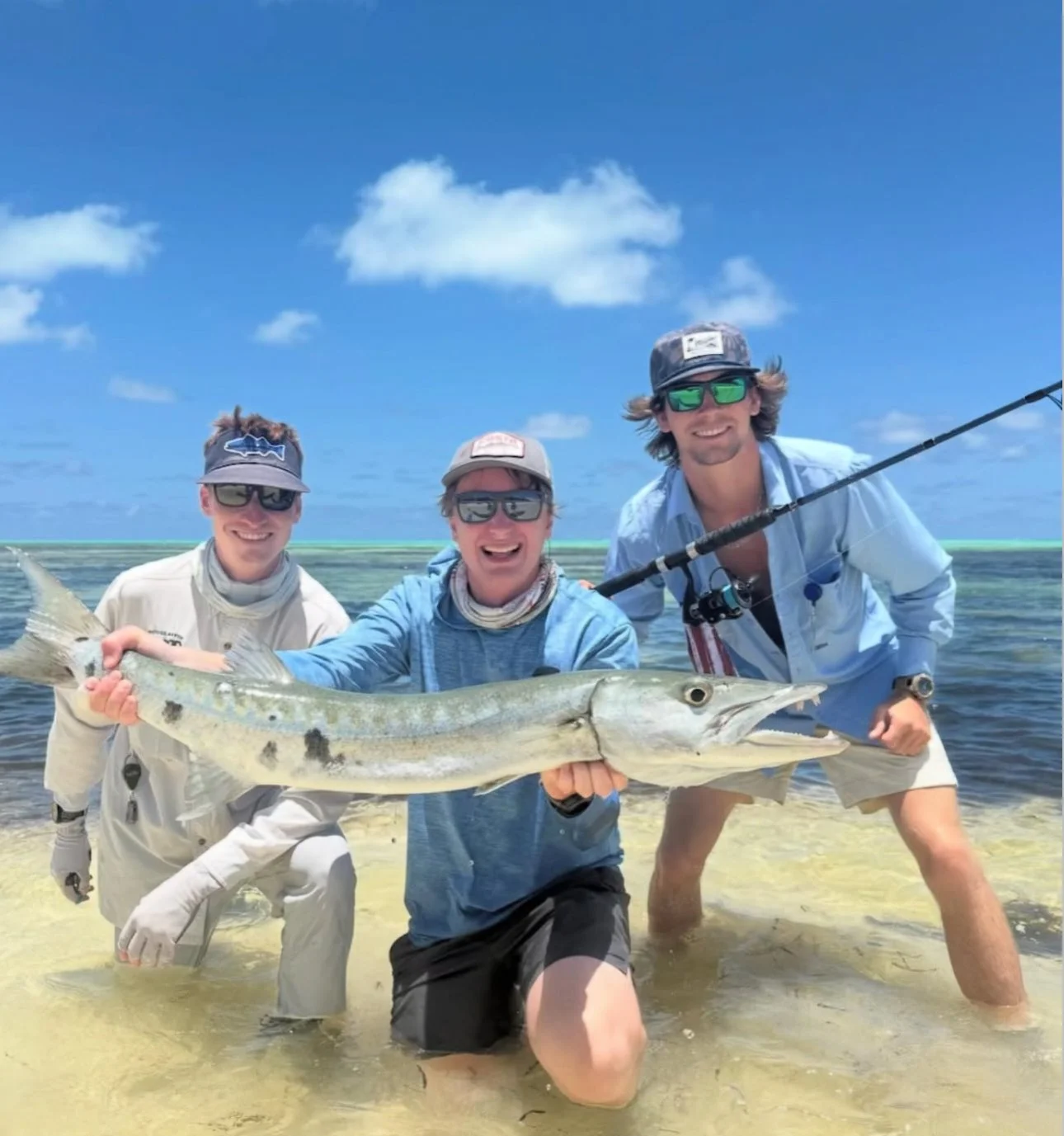 Three people standing in shallow water at the beach, holding a large bonefish and smiling. The middle person is holding the bonefish horizontally. The background shows a blue sky with some clouds and a distant shoreline.