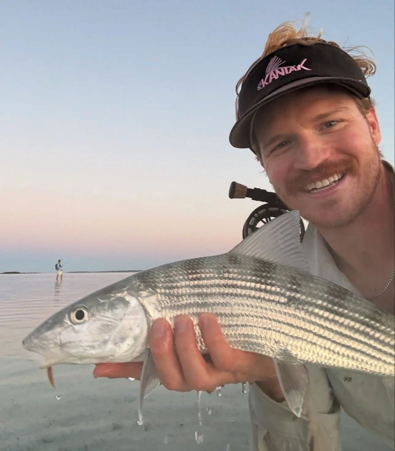 A man smiling and holding a bonefish in a shallow body of water during sunset, with another person fishing in the background.