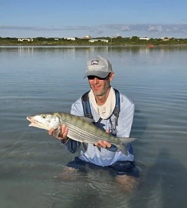 Man in waders holding a large bonefish in shallow water, with a calm lake and distant buildings in the background.