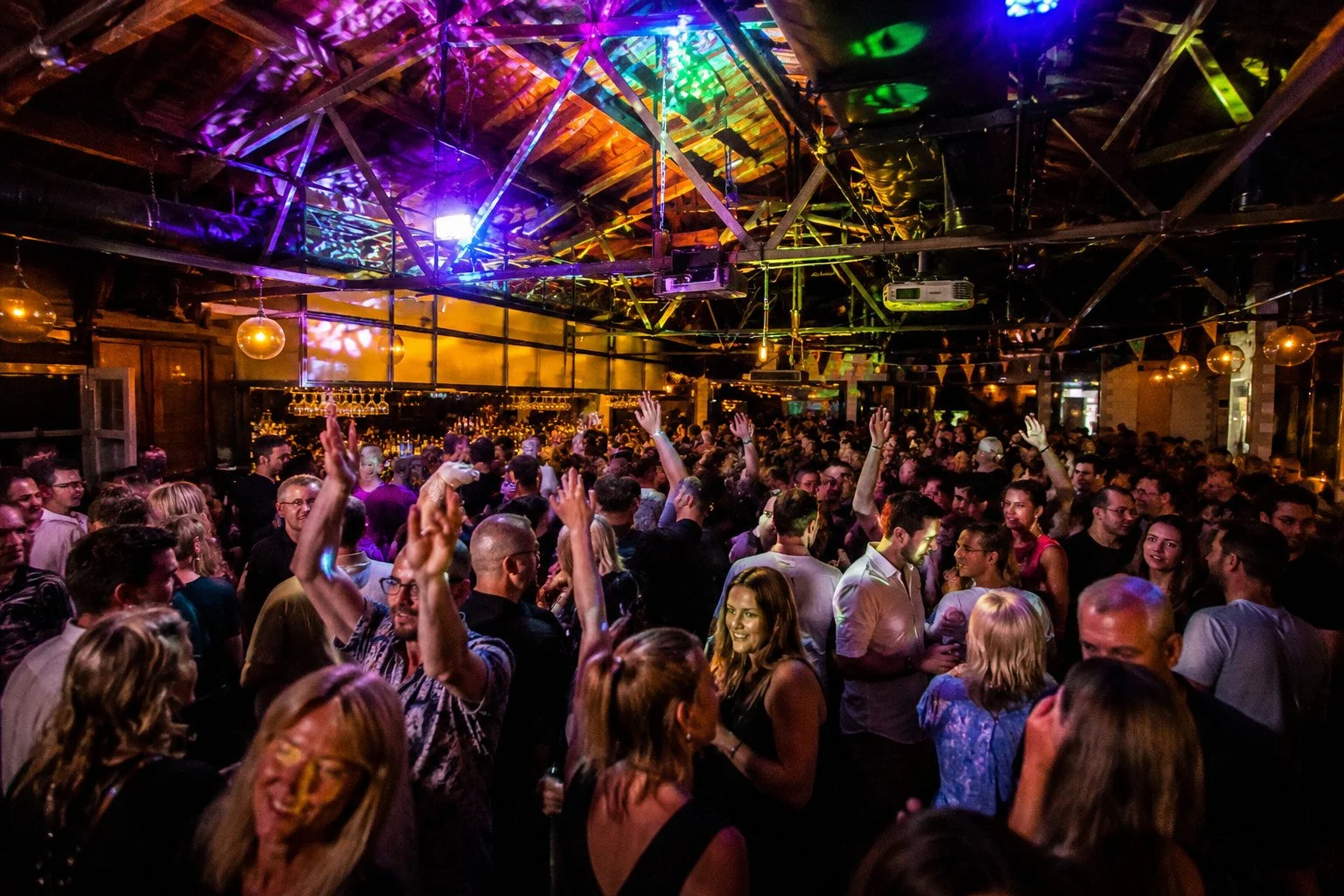 Crowd of people dancing and socializing in an indoor nightclub with colorful lighting and exposed wooden beams.