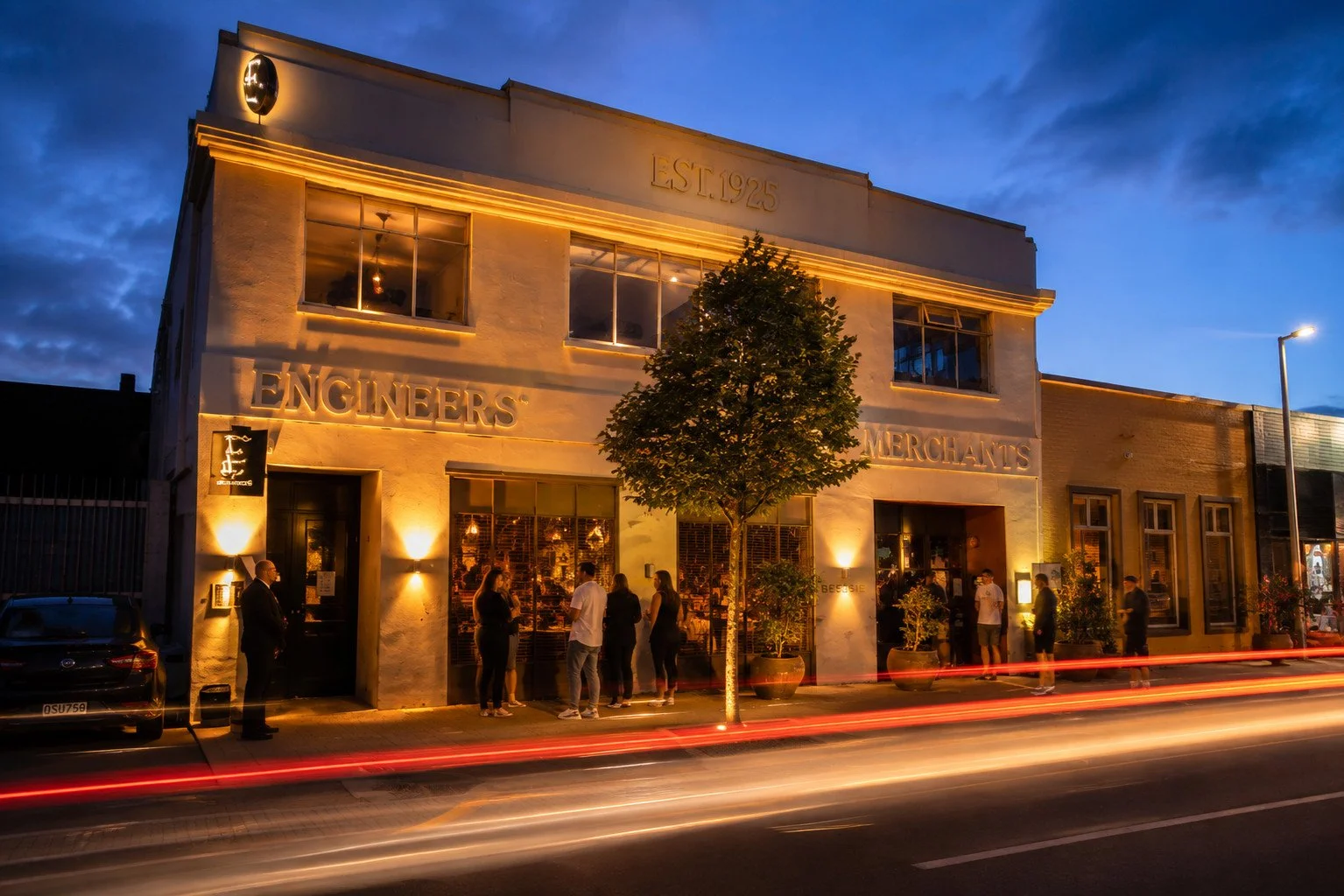 The facade of a historic building with the inscription 'EST. 1925' and words 'ENGINEERS' and 'MERCHANTS' on it, illuminated with warm lights at dusk, with people standing outside and moving cars creating light trails in front.