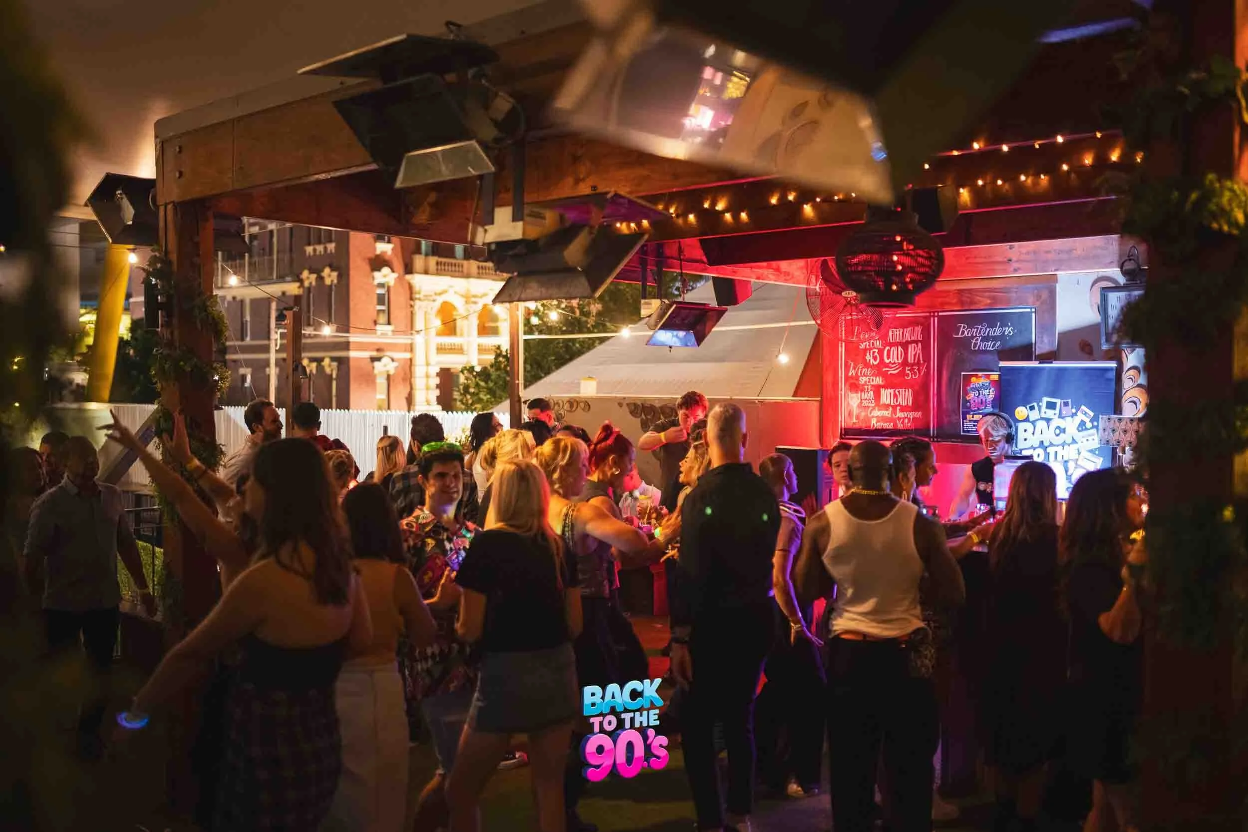 A lively outdoor party at night with people dancing and socializing, decorated with string lights and signs referencing the 80s and 90s.