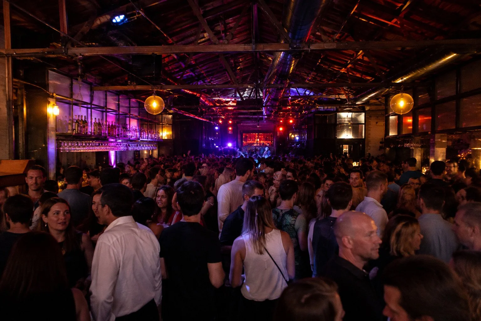 A crowded indoor concert or club with people standing and talking, colorful stage lighting, and a stage at the far end with performers.