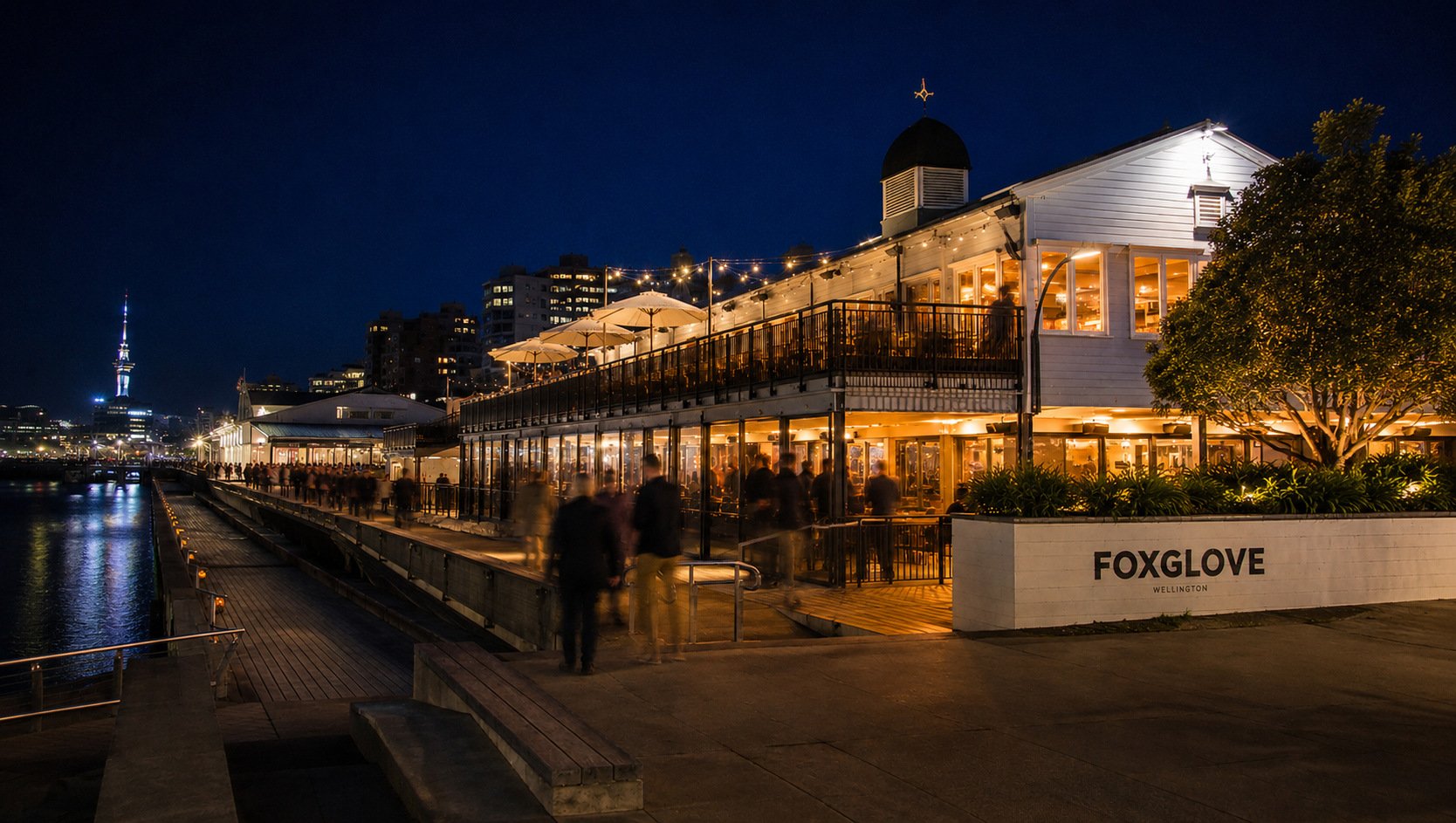 Nighttime scene of the Foxglove restaurant in Wellington, illuminated with warm lights, with people walking by along the waterfront promenade, and city buildings and the Sky Tower visible in the distance.