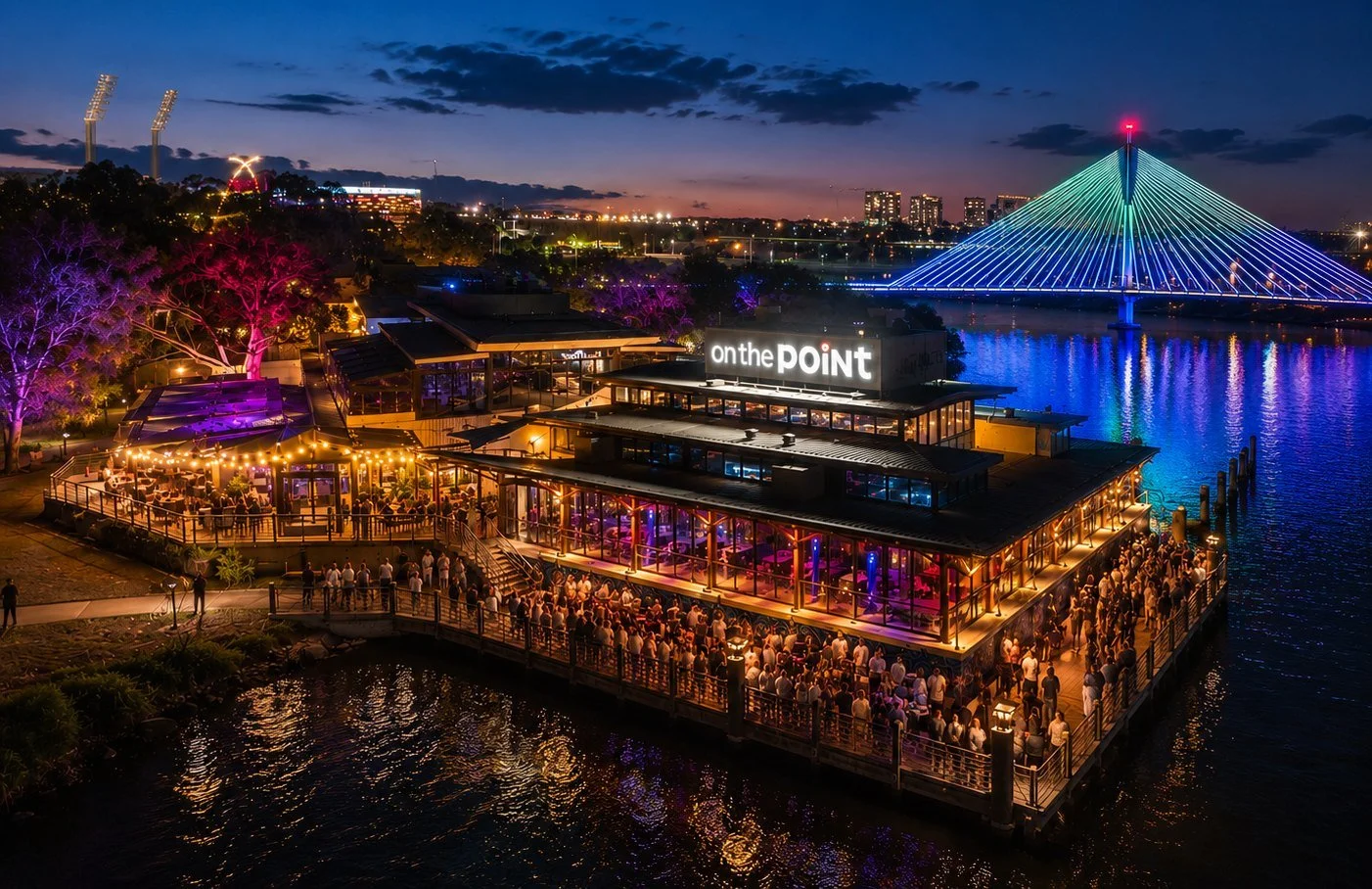 Night view of a waterfront restaurant named 'on the point' with many people dining, colorful trees, and a lit-up bridge over the water in a city skyline.