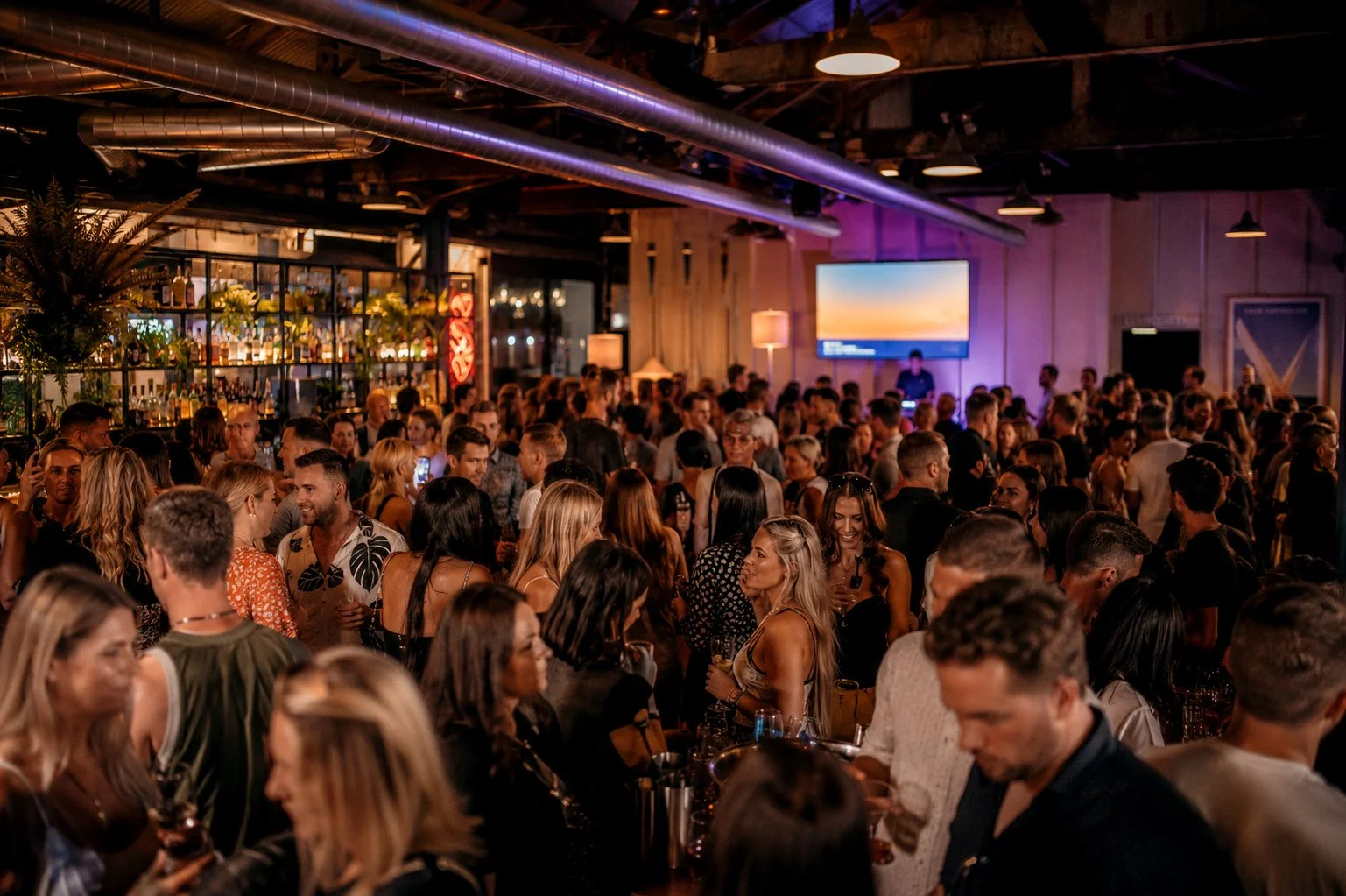 A crowded bar or nightclub with many people socializing, drinking, and enjoying events. The setting has warm lighting, a bar area with bottles visible, and a large screen displaying a sunset scene.