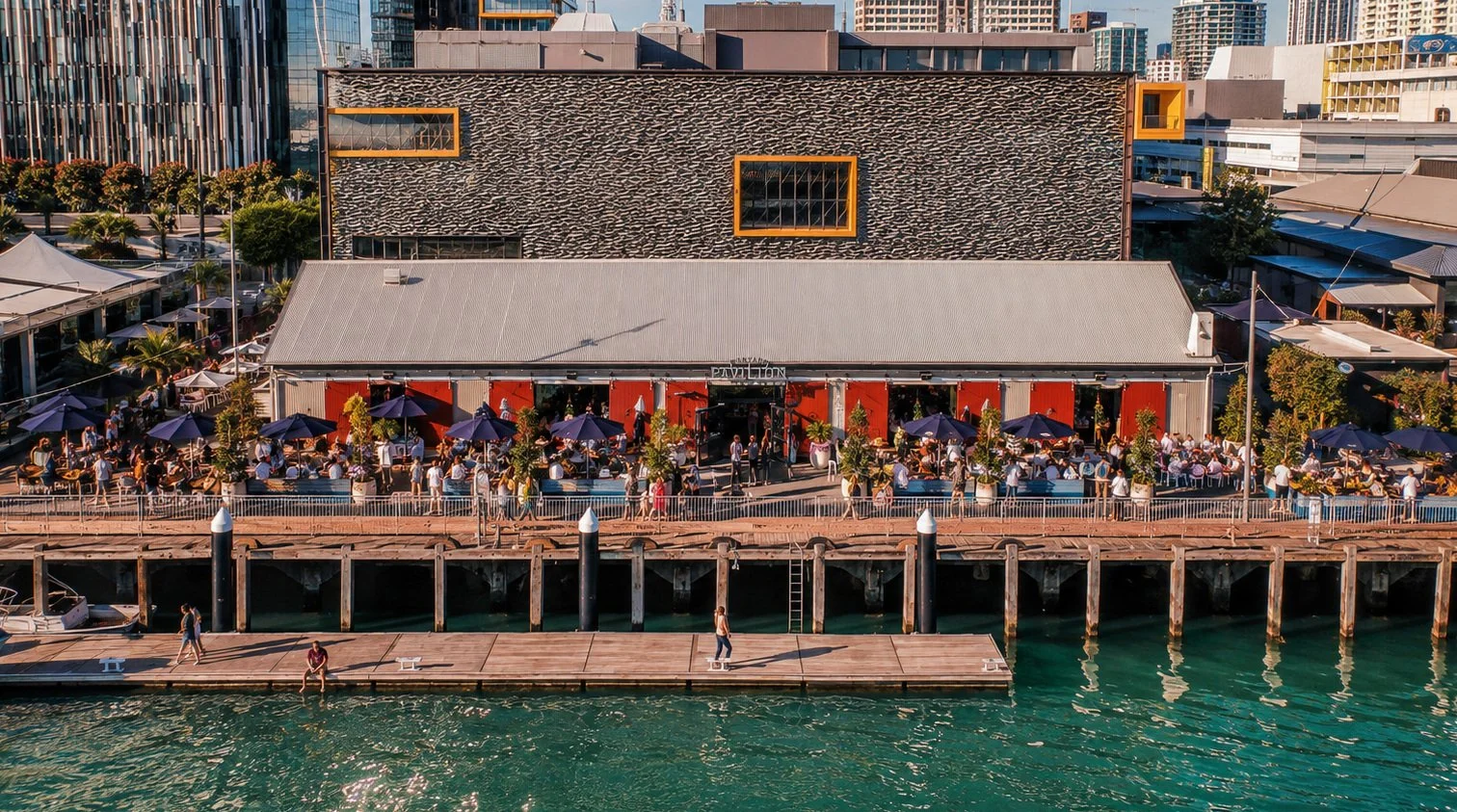 A waterfront building with a patio filled with people dining under purple umbrellas, adjacent to a wooden dock on a sunny day, with city buildings in the background.