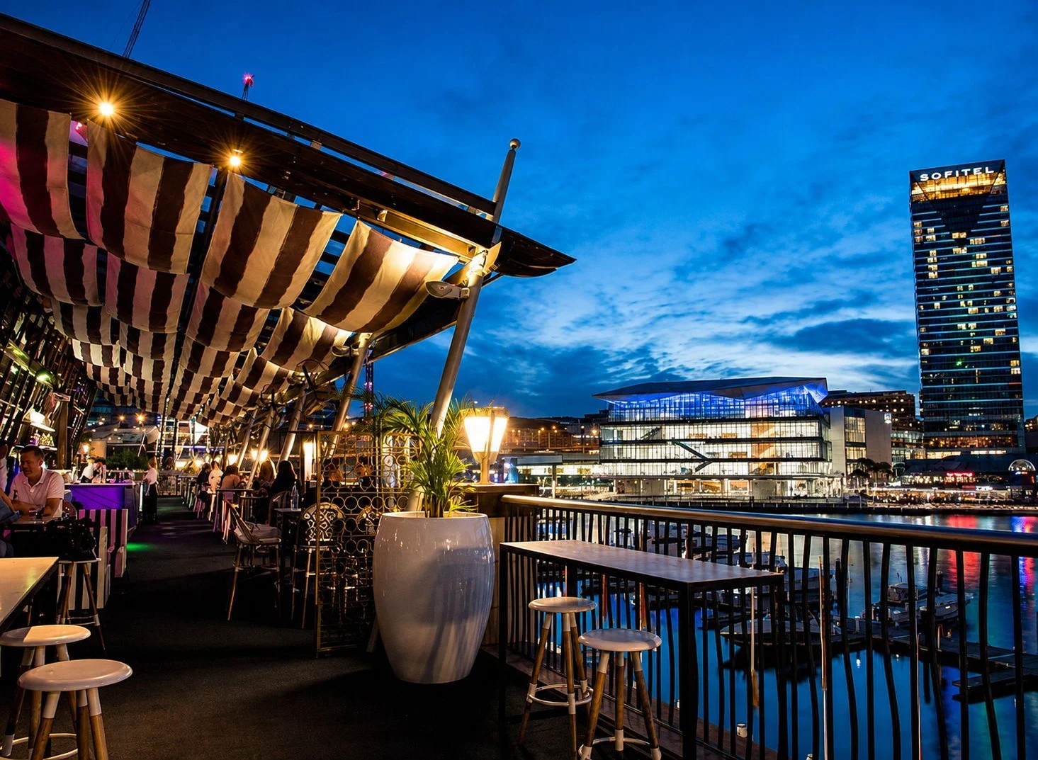 A rooftop bar with a view of a city skyline at dusk, featuring a large building with the 'Sofitel' sign, outdoor seating, and decorative lighting.