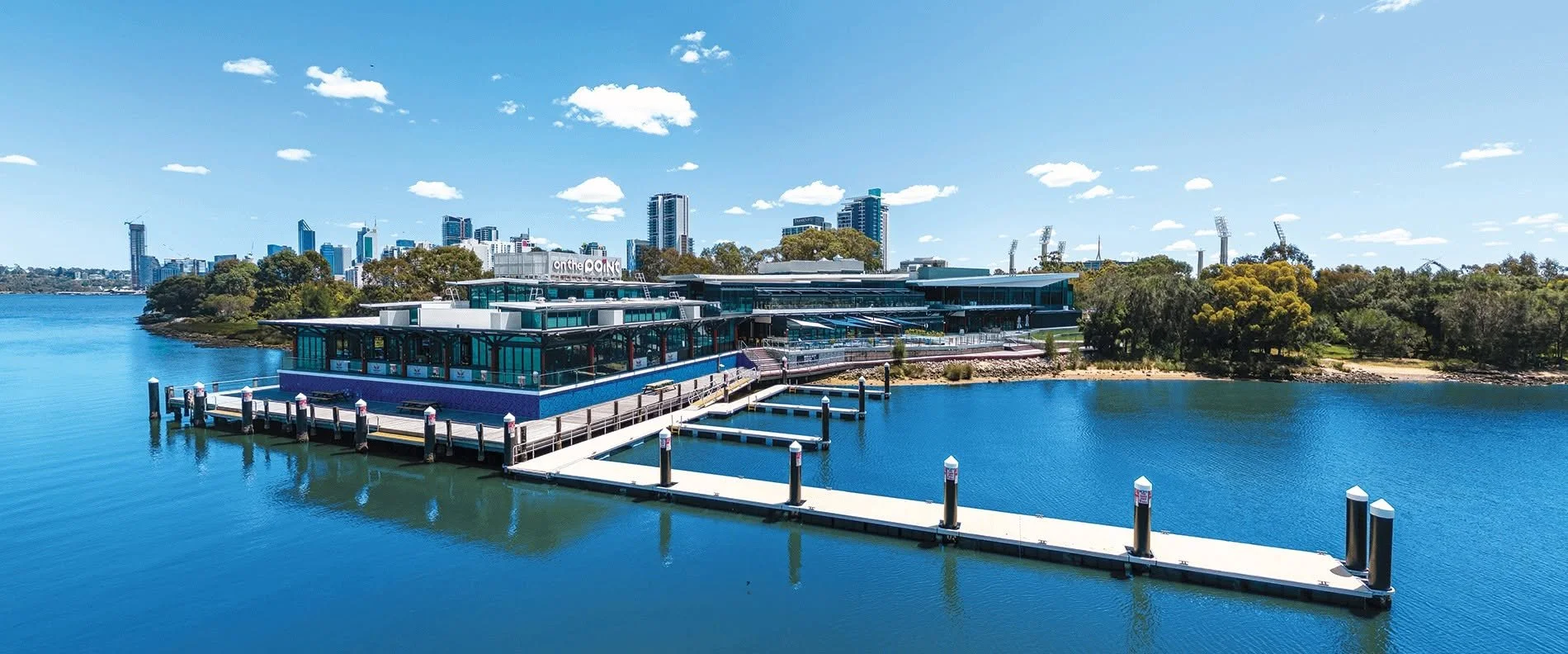 Modern waterfront building with glass windows and outdoor seating, situated on a pier extending into calm blue water, with trees and a city skyline in the background under a partly cloudy sky.