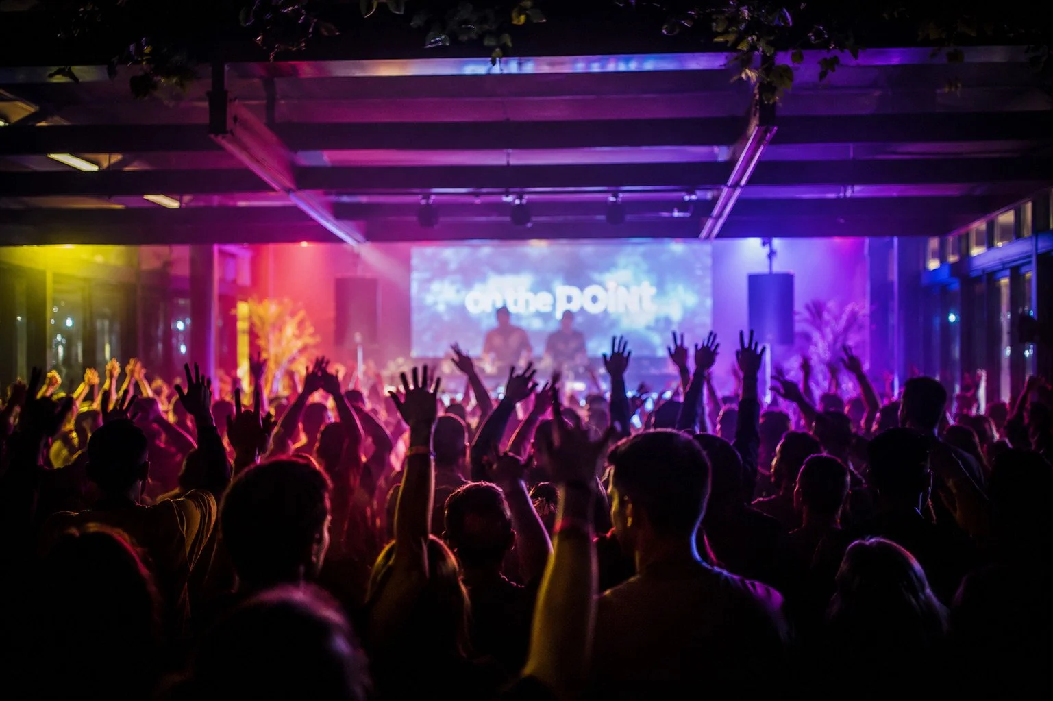 Crowd of people at a concert or event with their hands raised, stage with performers and a large screen displaying 'on the point' in the background, colorful lighting in purple, pink, and yellow.
