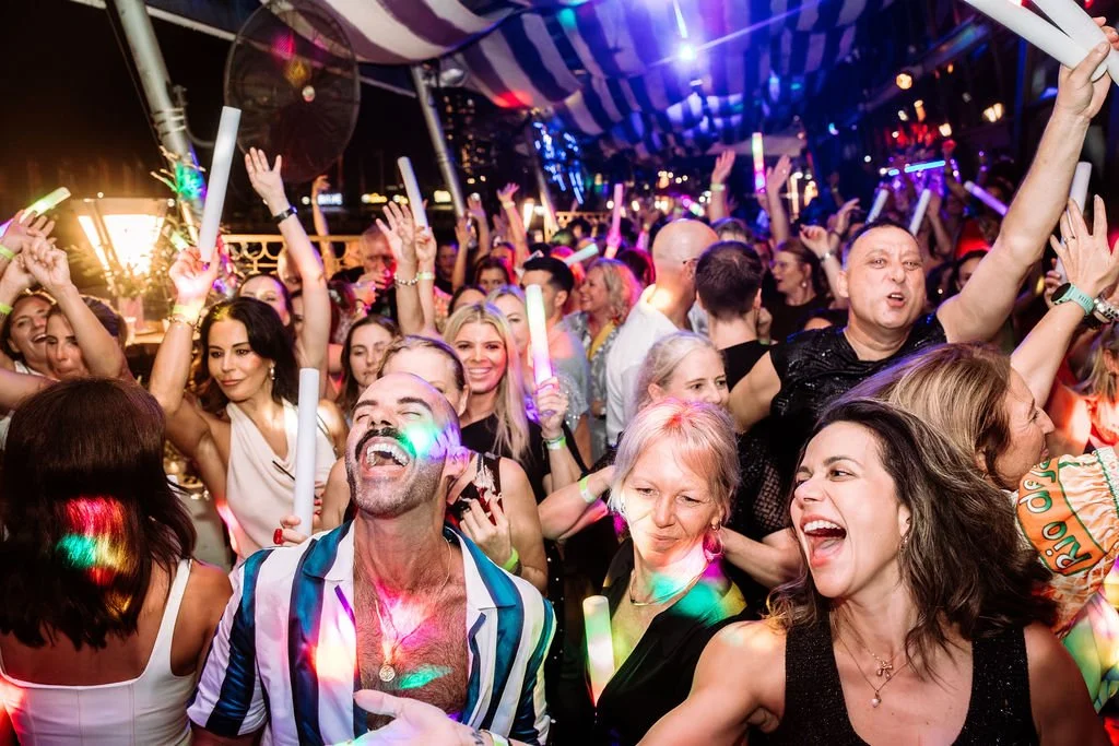 A lively crowd of people dancing and celebrating at a party or concert with colorful lights and glow sticks.