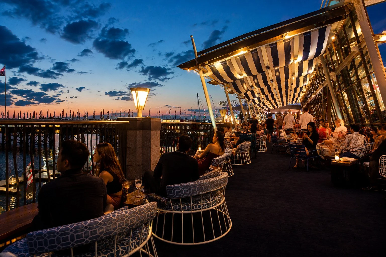 Outdoor rooftop bar at sunset with people dining and socializing, overlooking a body of water with sailboats, illuminated lamps, and a partly cloudy sky.