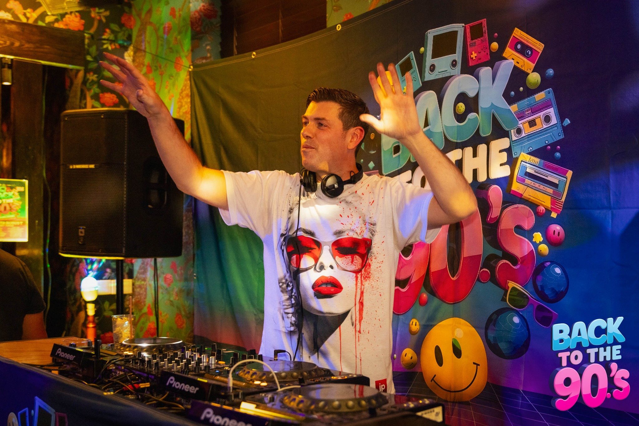 DJ with headphones wearing a white t-shirt featuring a woman's face with red makeup and sunglasses, raising his hands in a colorful, 90s-themed party setting.