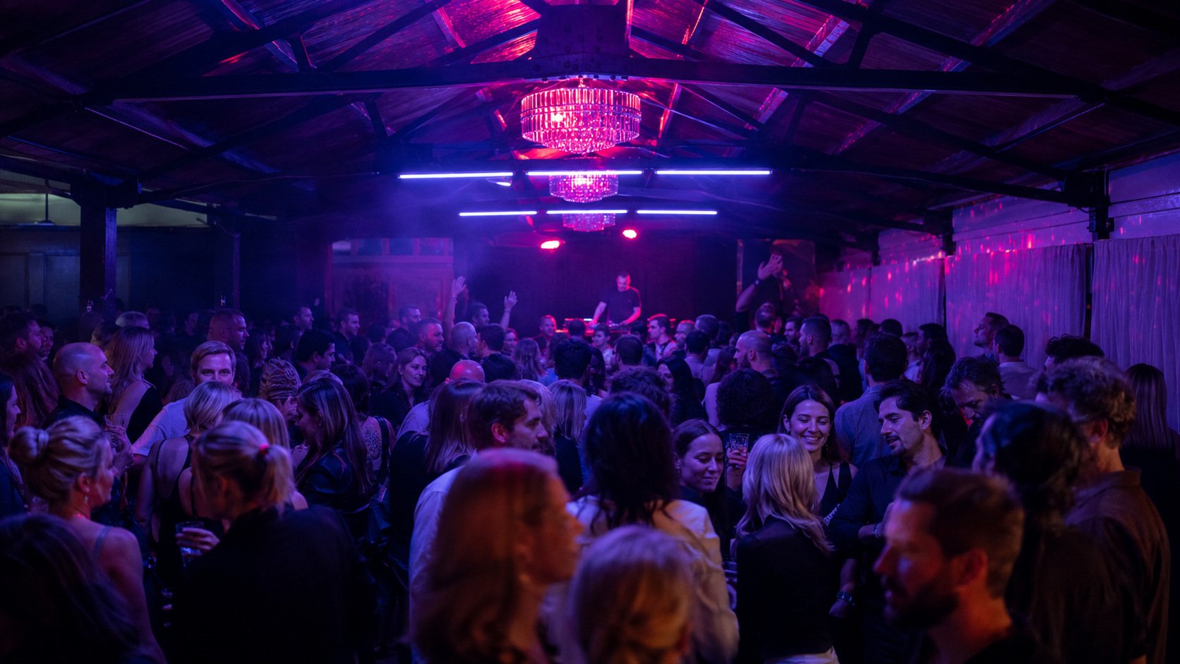 Crowd of people dancing and socializing at a nightclub with purple and pink lighting, a DJ booth in the center, and chandeliers hanging from the ceiling.