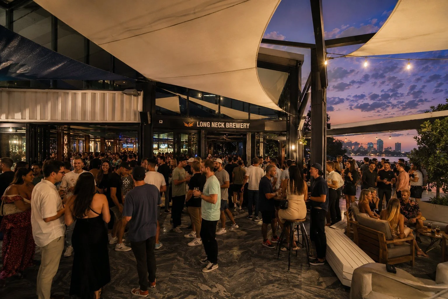 A crowded outdoor bar scene at Long Neck Brewery during sunset, with groups of people socializing, some sitting on benches, others standing and drinking, overlooking a city skyline and river.