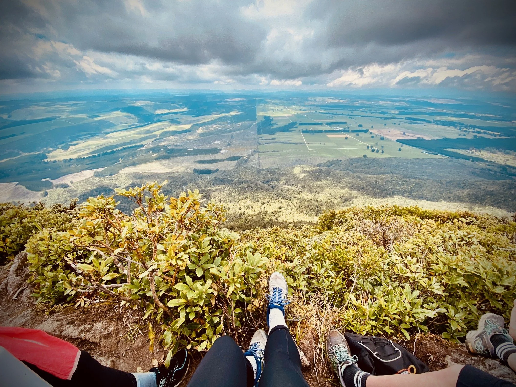 Three people sitting on a mountain ledge overlooking a valley with fields, forests, and clouds in the sky.