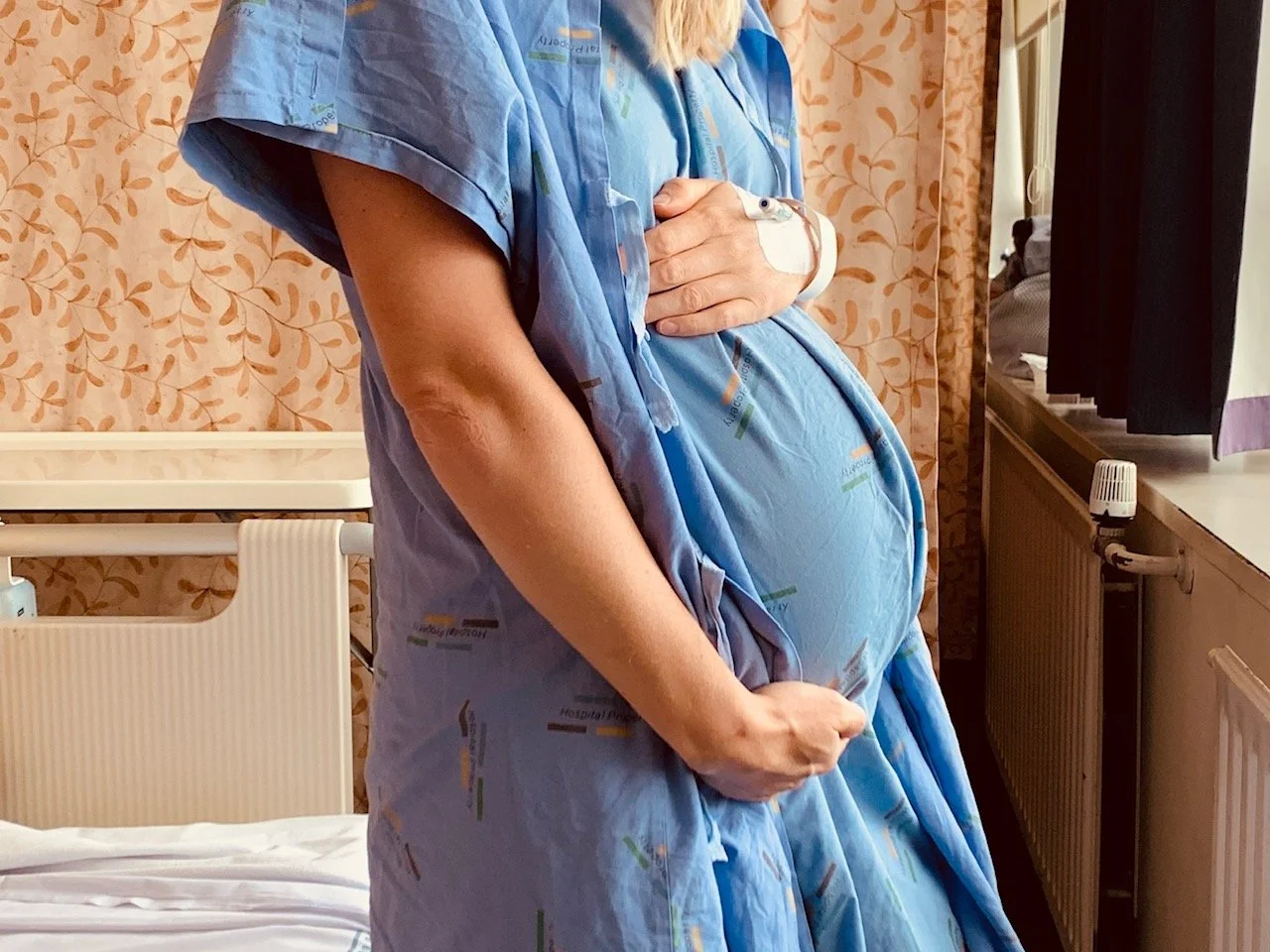 A pregnant woman in hospital gown standing beside a bed, holding her belly with one hand and placing her other hand over her chest.
